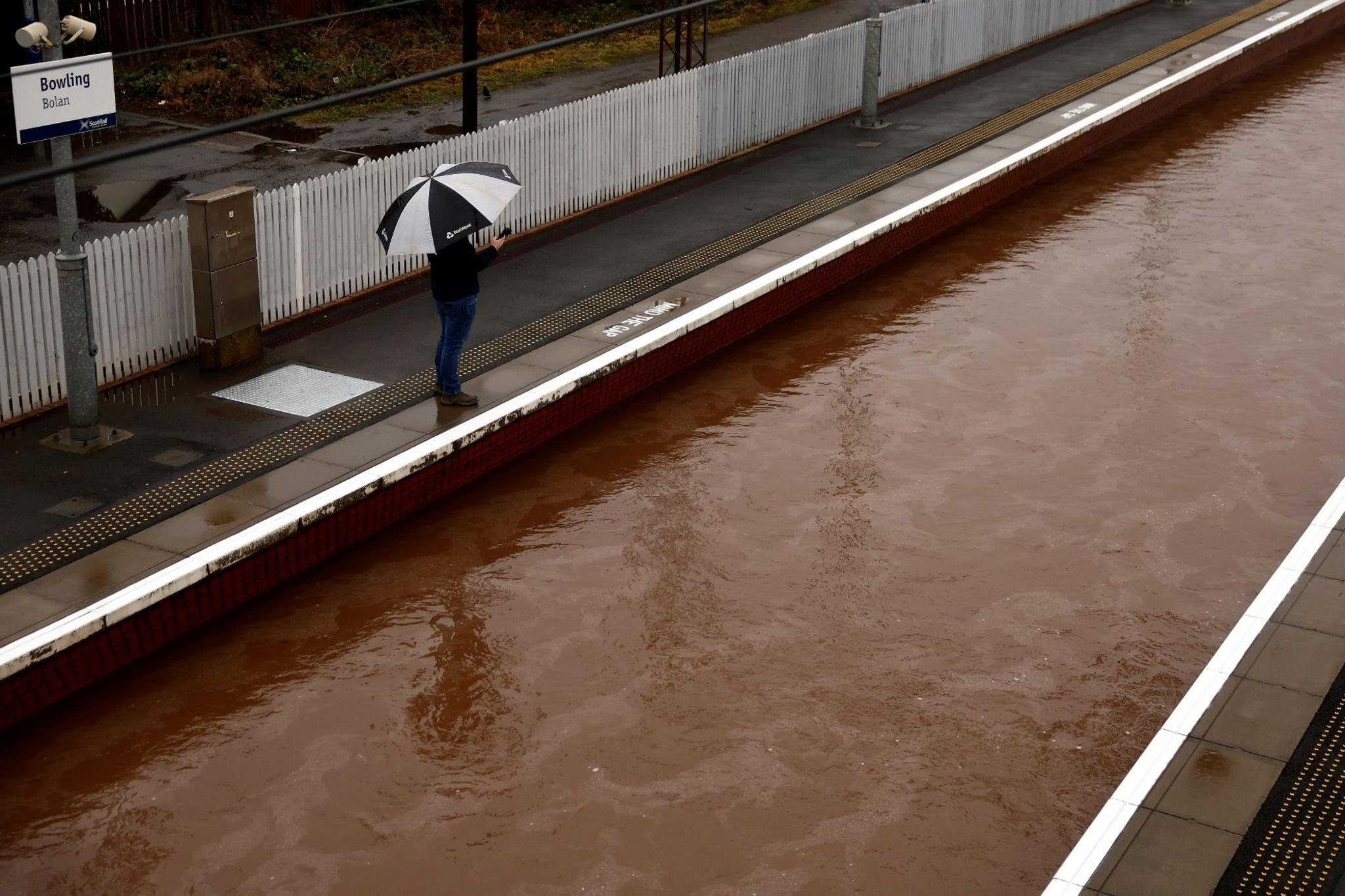 A man with a black and white umbrella stands at the edge of a railway platform with the entire track covered with flood waters.