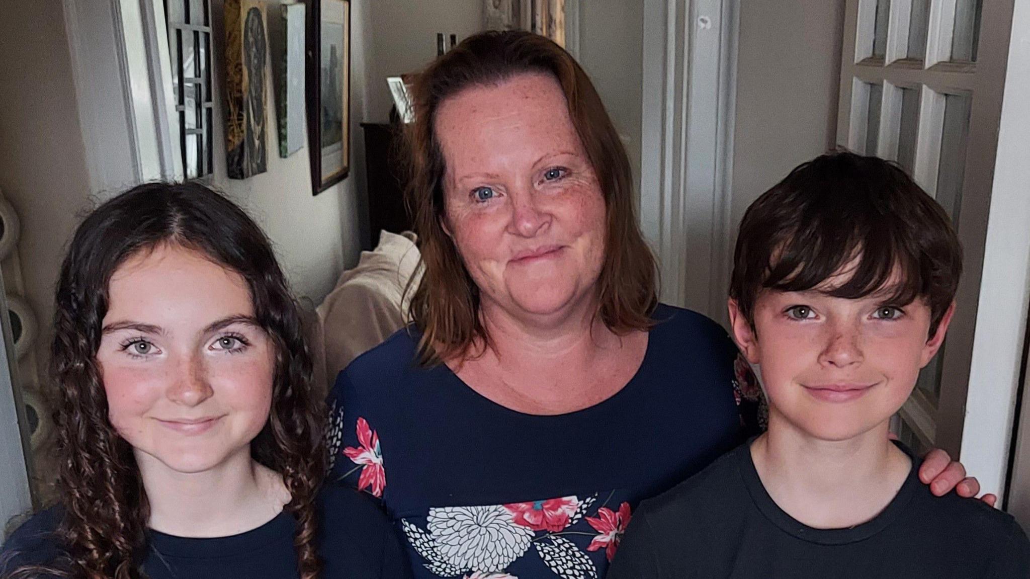 Liza Ellen and her two children smile directly at the camera, photographed standing in their house. The children are wearing dark T-shirts and Liza has a top with a flowery pattern.