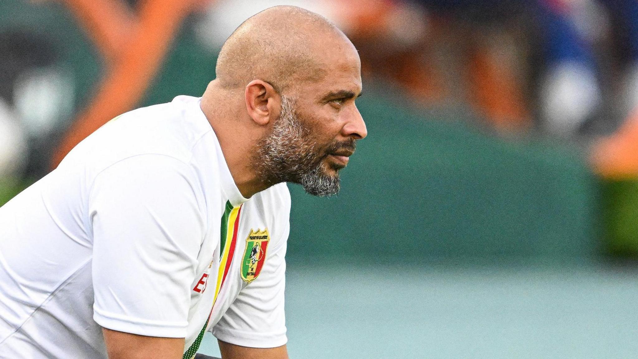 Eric Chelle, wearing a white Mali training top, leans forward with an expression of concentration