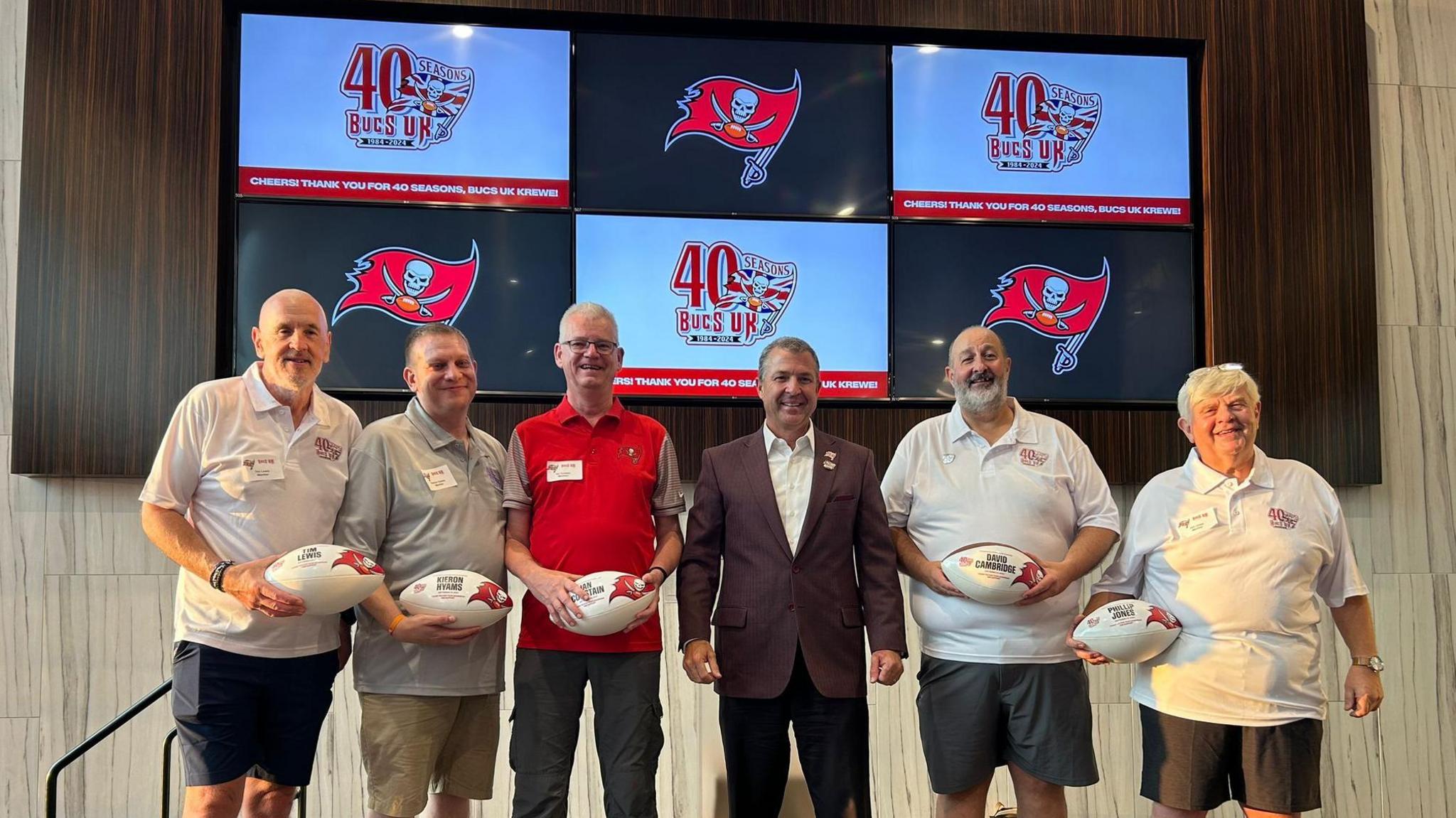 Five members receiving named game balls on stage at the club's 40th birthday meal in Tampa, which was organised by the Buccaneers. Brian Ford is stood centre and screens with the clubs logo and team logo are behind them.