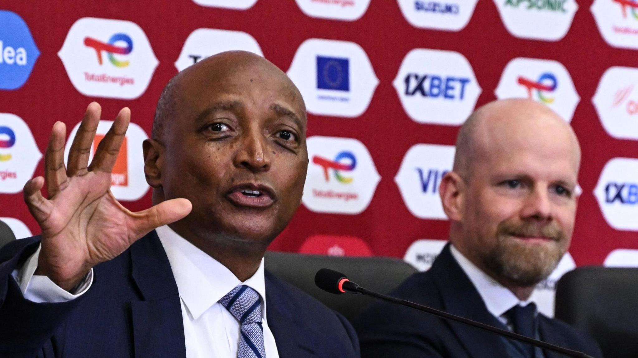 Patrice Motsepe seen from chest up gesticulates in the direction of the camera with the open palm of his right hand as he speaks into a microphone at a press conference. Motsepe is on the left-hand side of the image, which is the near side, with Mattias Grafstrom of Fifa seated on the far side of his, which is the right-hand side of the picture. Both men are wearing suits