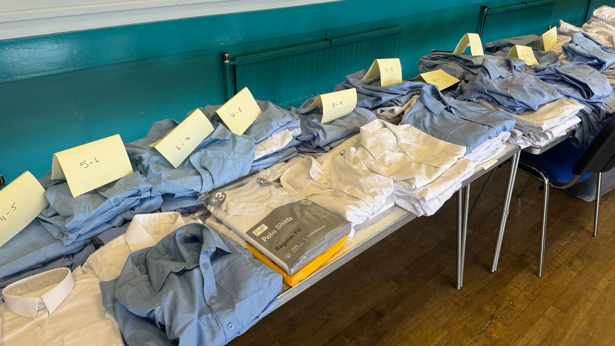 Blue and white school shirts are organised into piles and labelled by age on a table