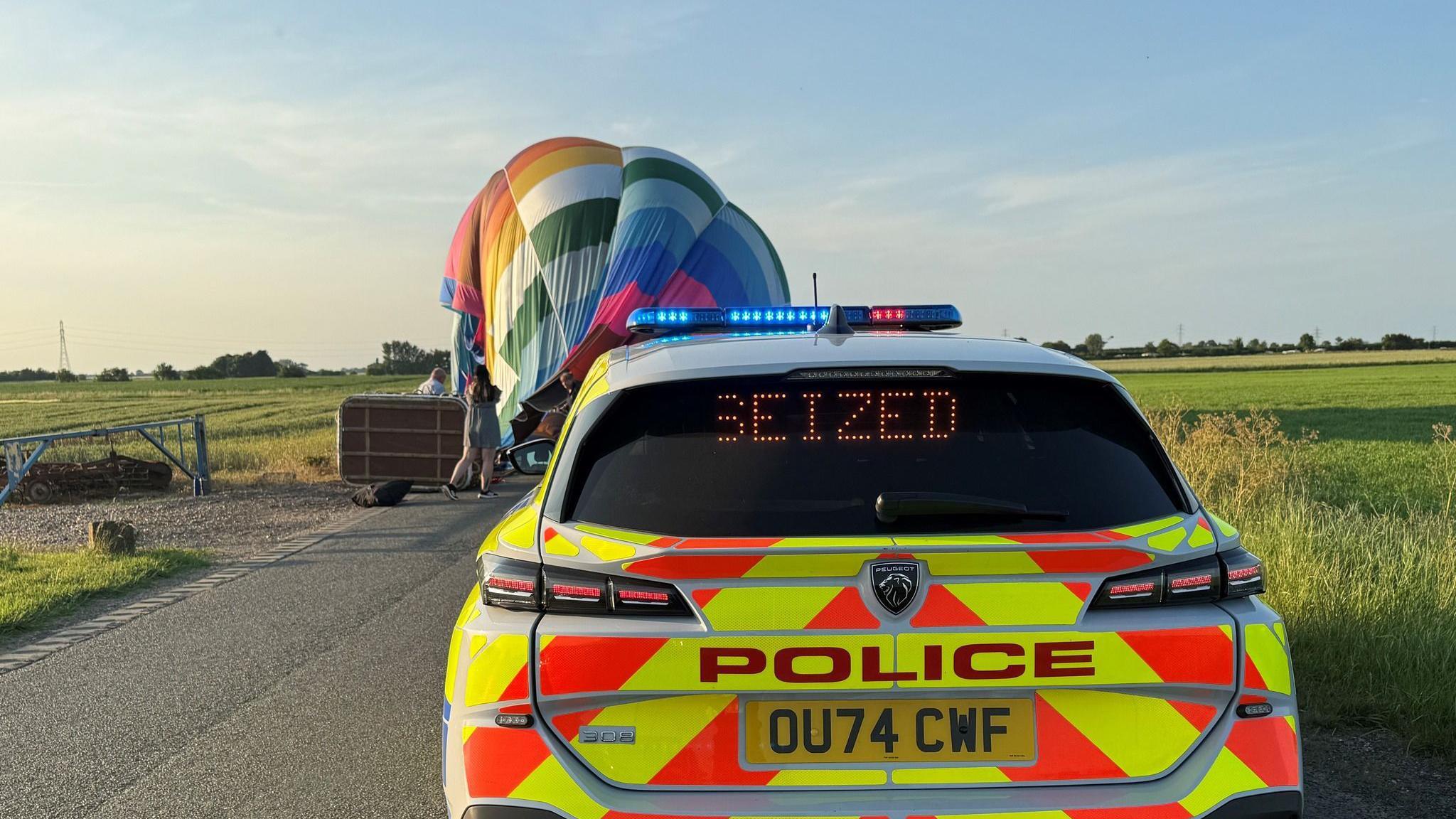 Brightly coloured deflated hot air balloon on side of rural road near fields. A police car is in front of the balloon.