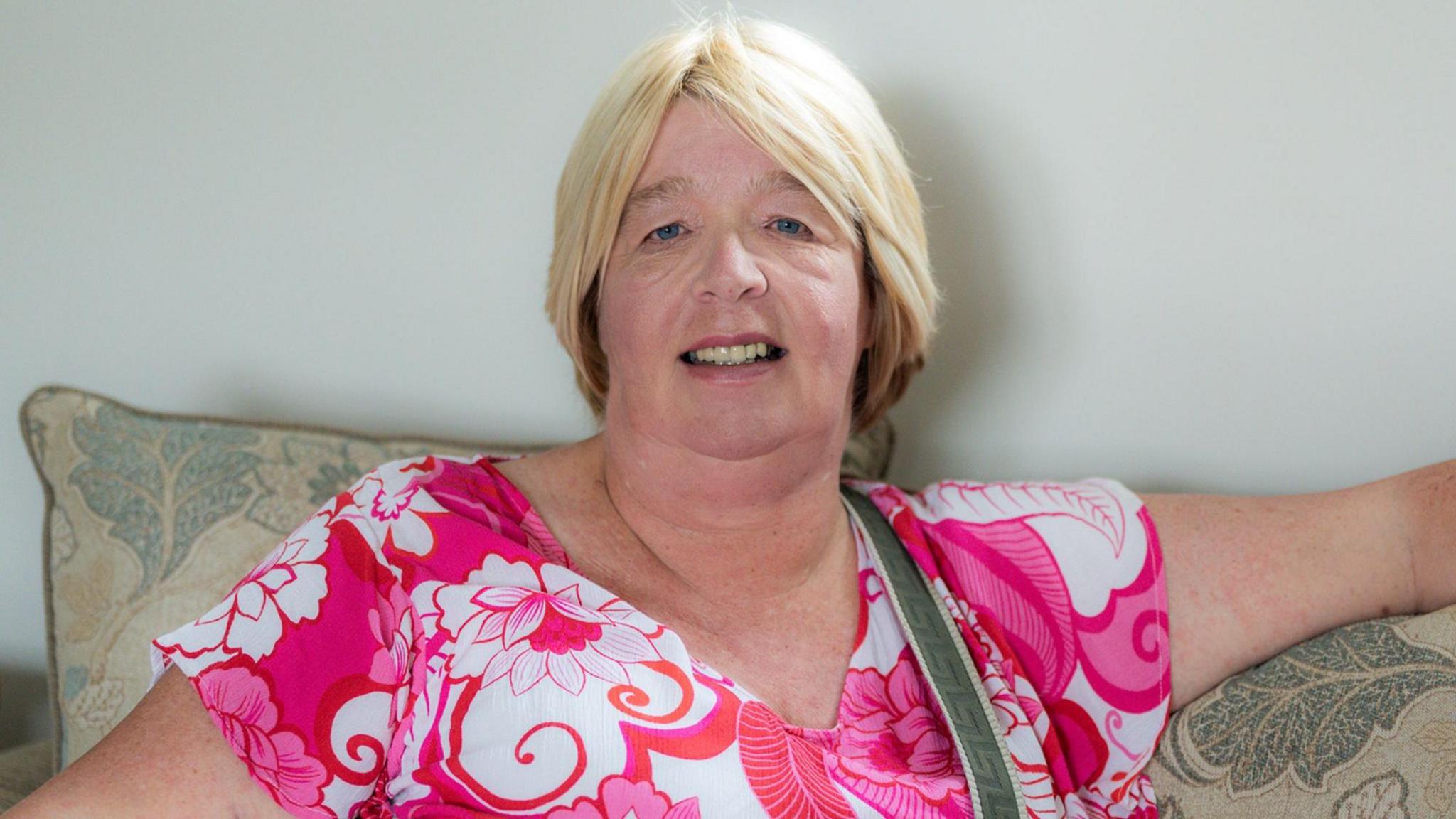 A woman with blonde hair is sat on a beige a green patterned sofa. She is wearing a pink and white flowery top.