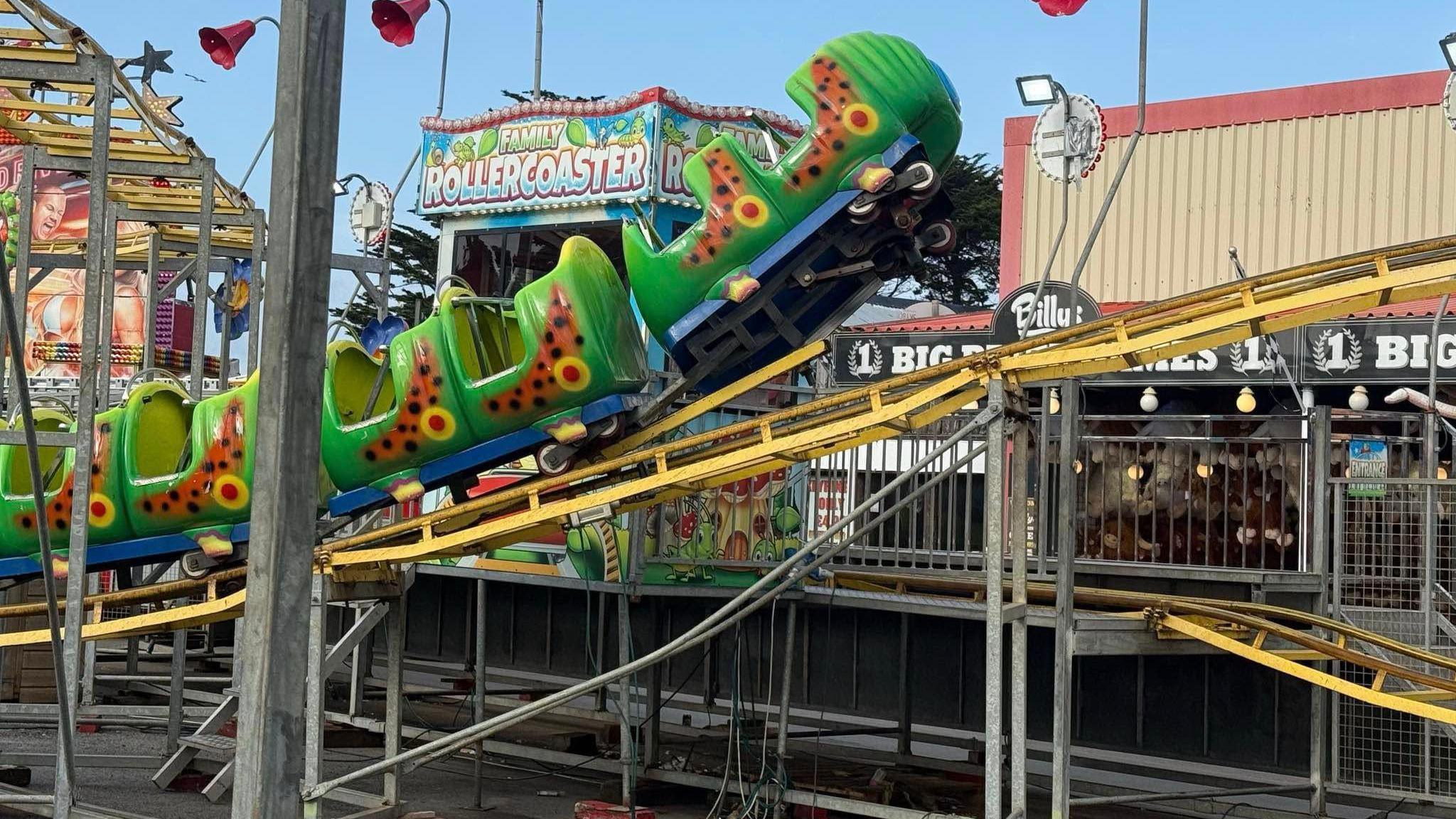 A side on view of a caterpillar themed rollercoaster where the front cart of the ride has come off the tracks. It is suspended in the air at the front as the yellow track goes out of the image. Behind there is a sign for a family rollercoaster