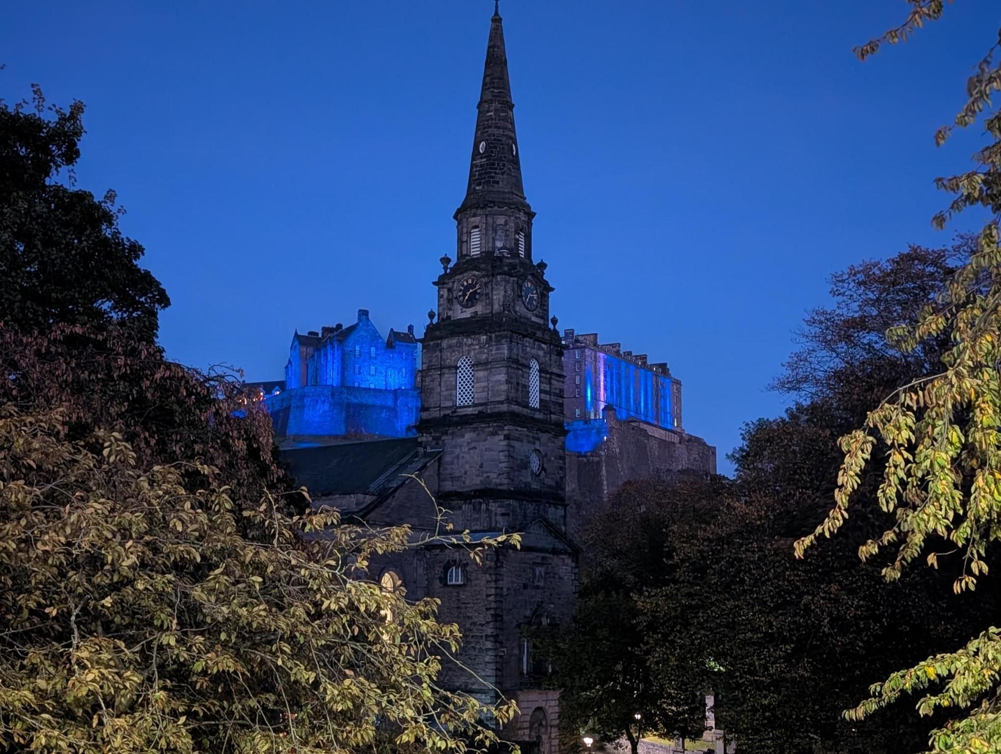 St Cuthbert church in Edinburgh in the foreground with Edinburgh Castle in the background, illuminated in blue lights below a clear, blue sky in the evening