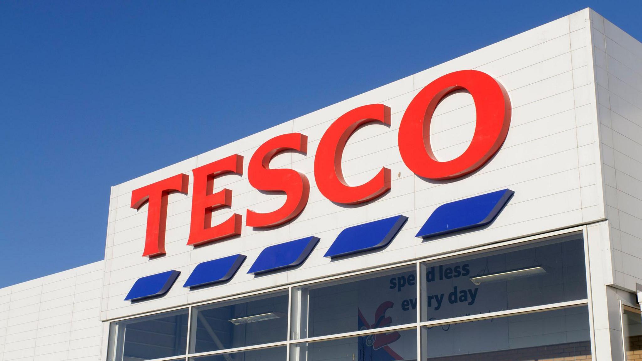 A Tesco logo on a white building. There is a blue sky above.