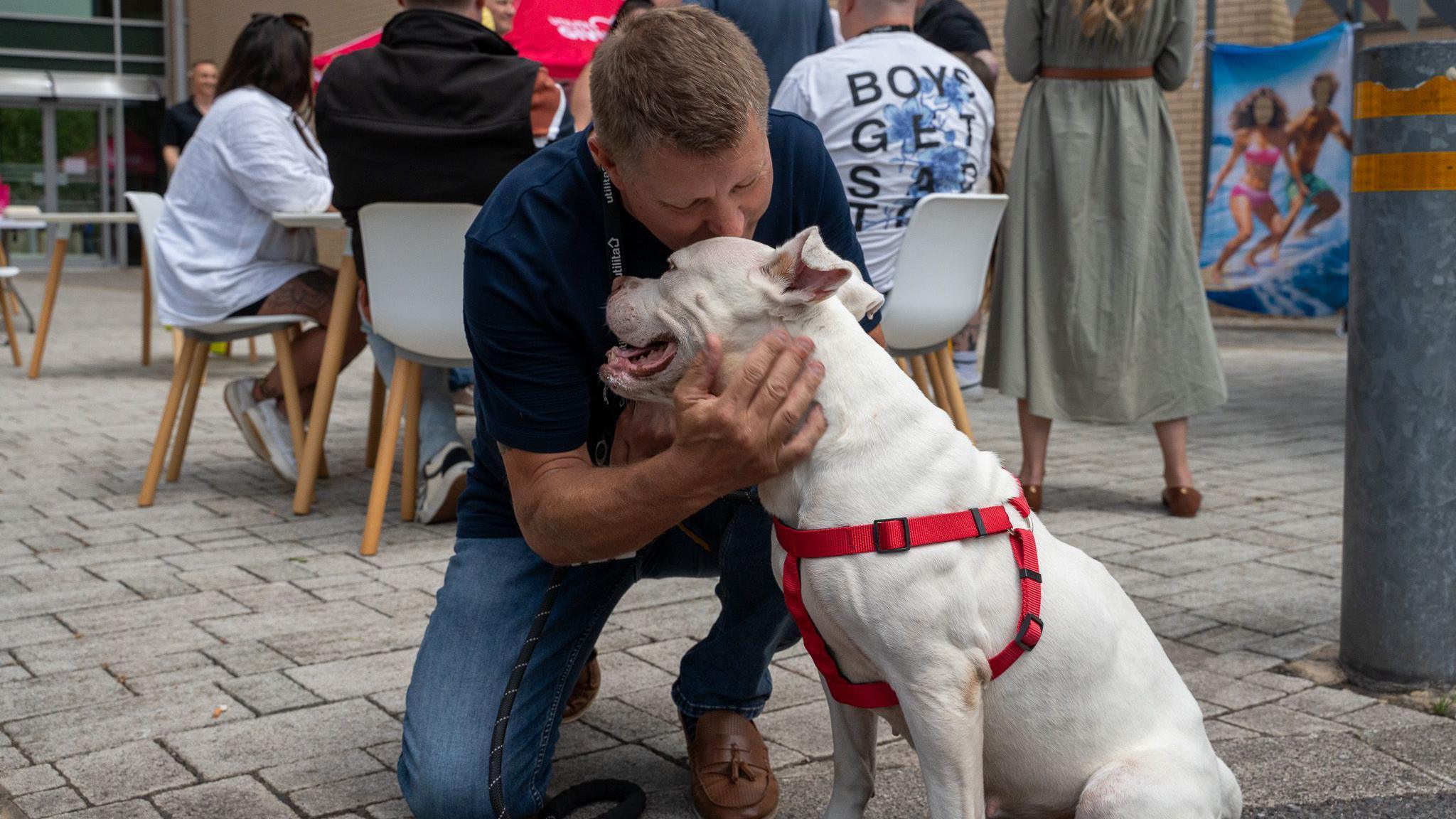 Tilly is sat looking away from the camera wearing her red harness. Shaun Dollamore is crouched next to her, he has his hands around her face affectionately and is leaning in to kiss the top of her head. Shaun has short dark blonde hair and wears a navy blue polo shirt with jeans and tan brown leather deck shows. In the background there are people sat around a table talking.
