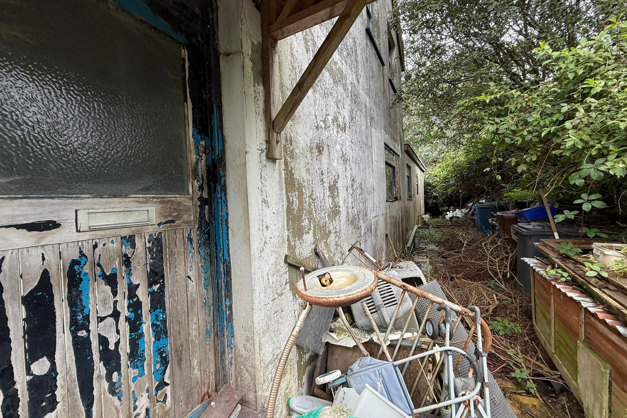 A close up of a pile of junk which is located just outside the house. A walker can be seen on top of the pile. Bins can be seen in the background in the overgrown garden. 