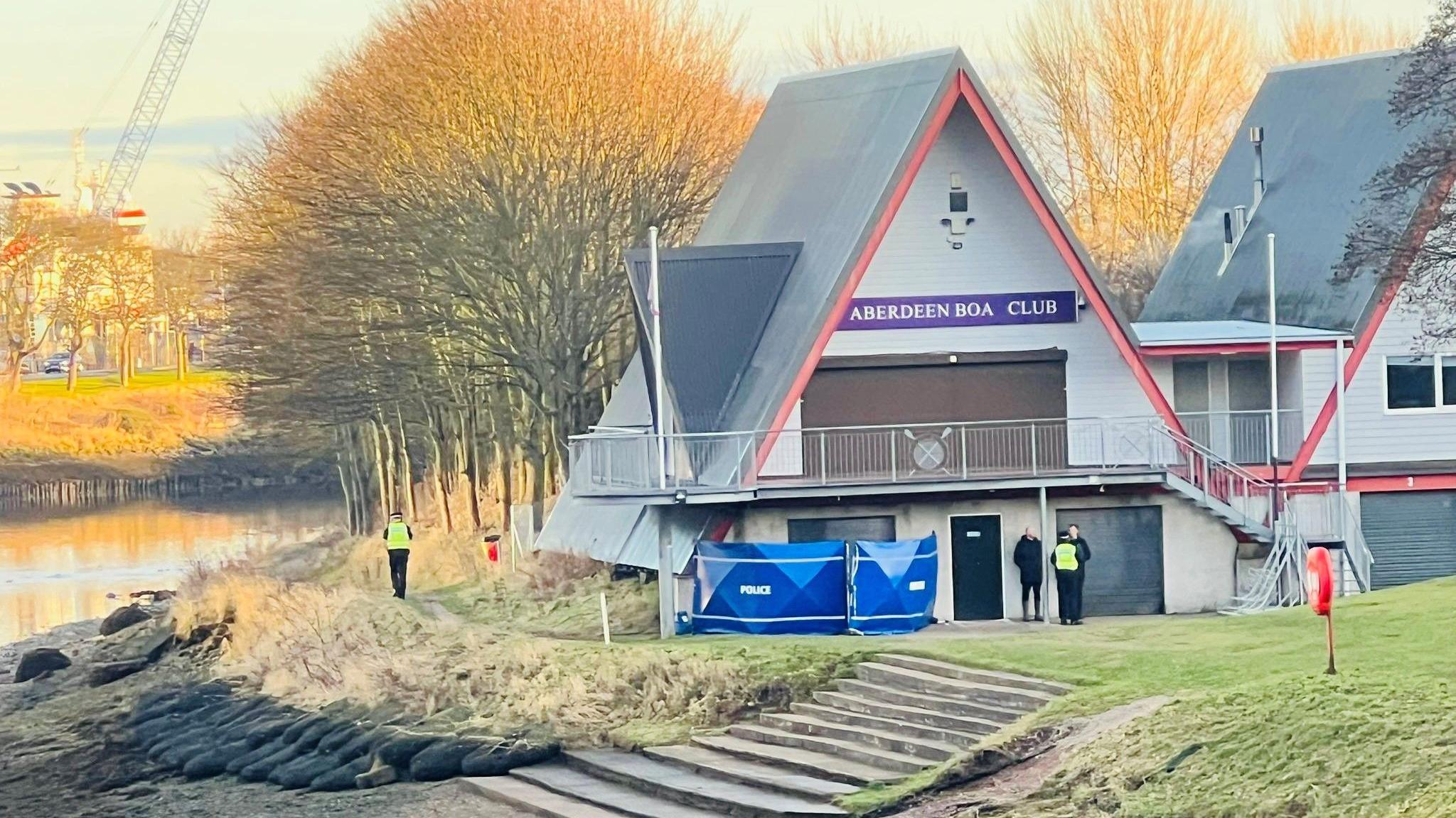 Blue police tents next to a river and Aberdeen Boat Club building.