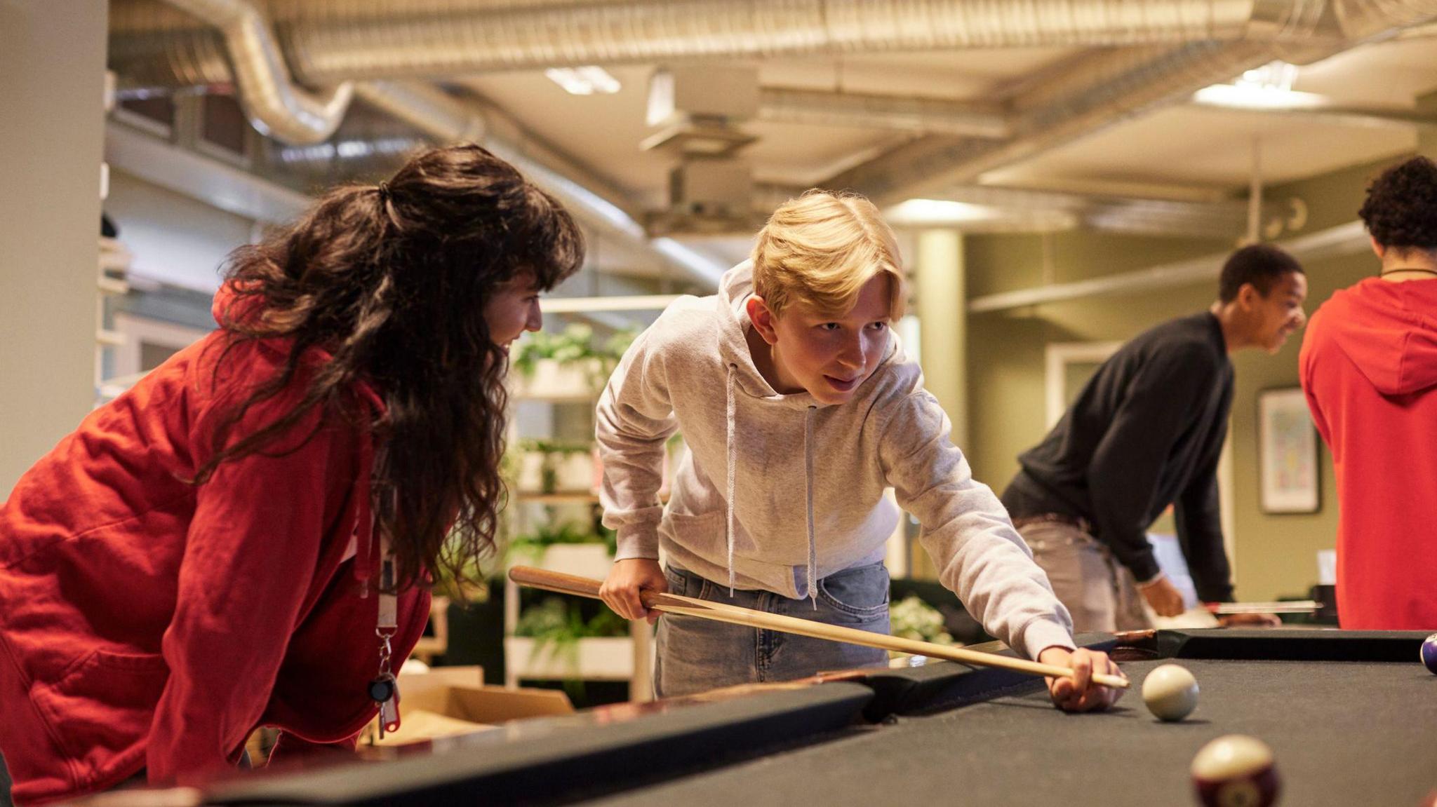 Female mentor assists a teenage boy aiming at a ball with pool cue stick.