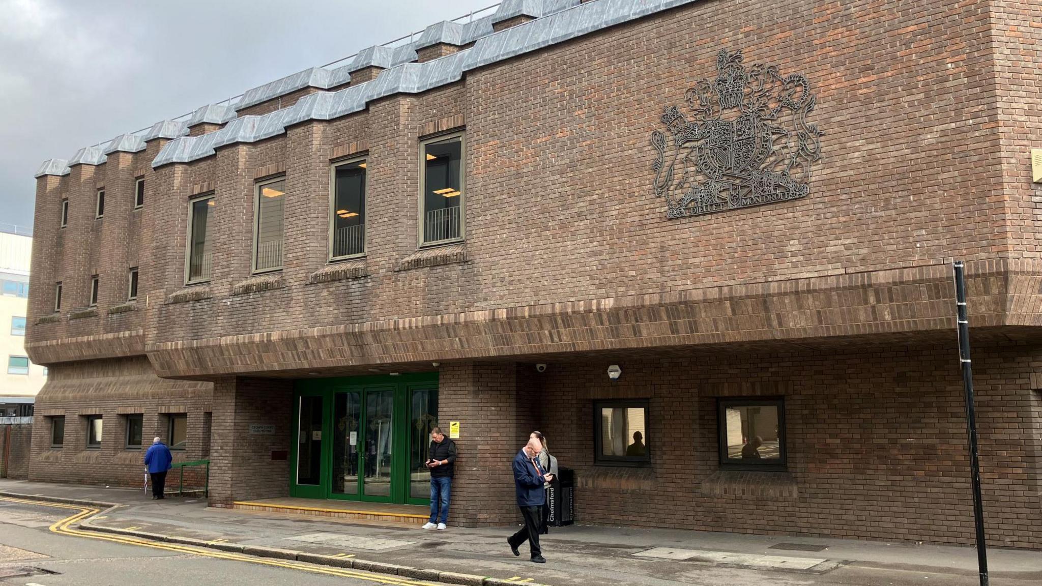 Chelmsford Crown Court, which is a red brick building that has green doors at its entrance. There are three people milling about outside the building.