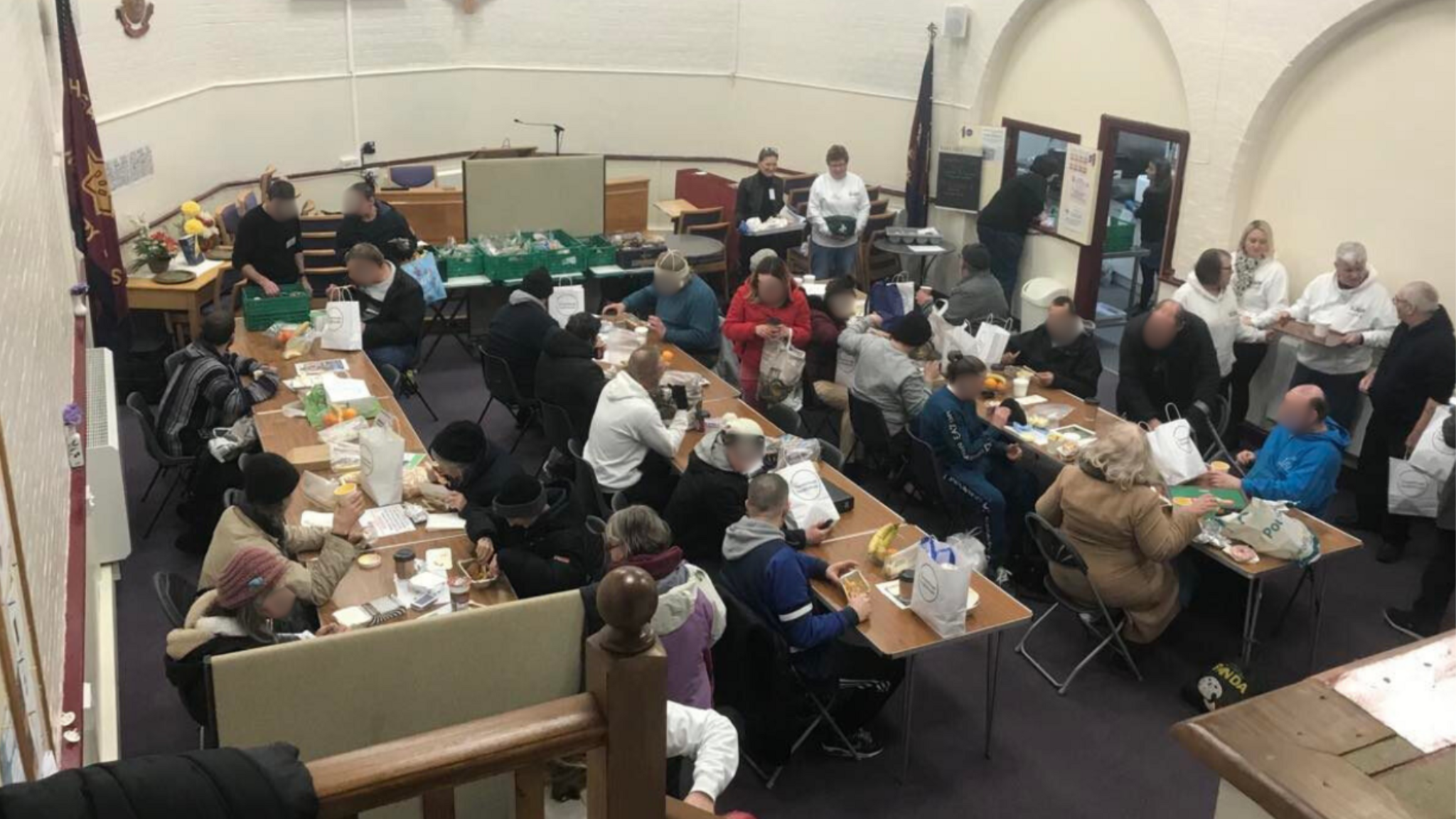 A crowd of people sat around tables eating with people serving them food on the edges of the room