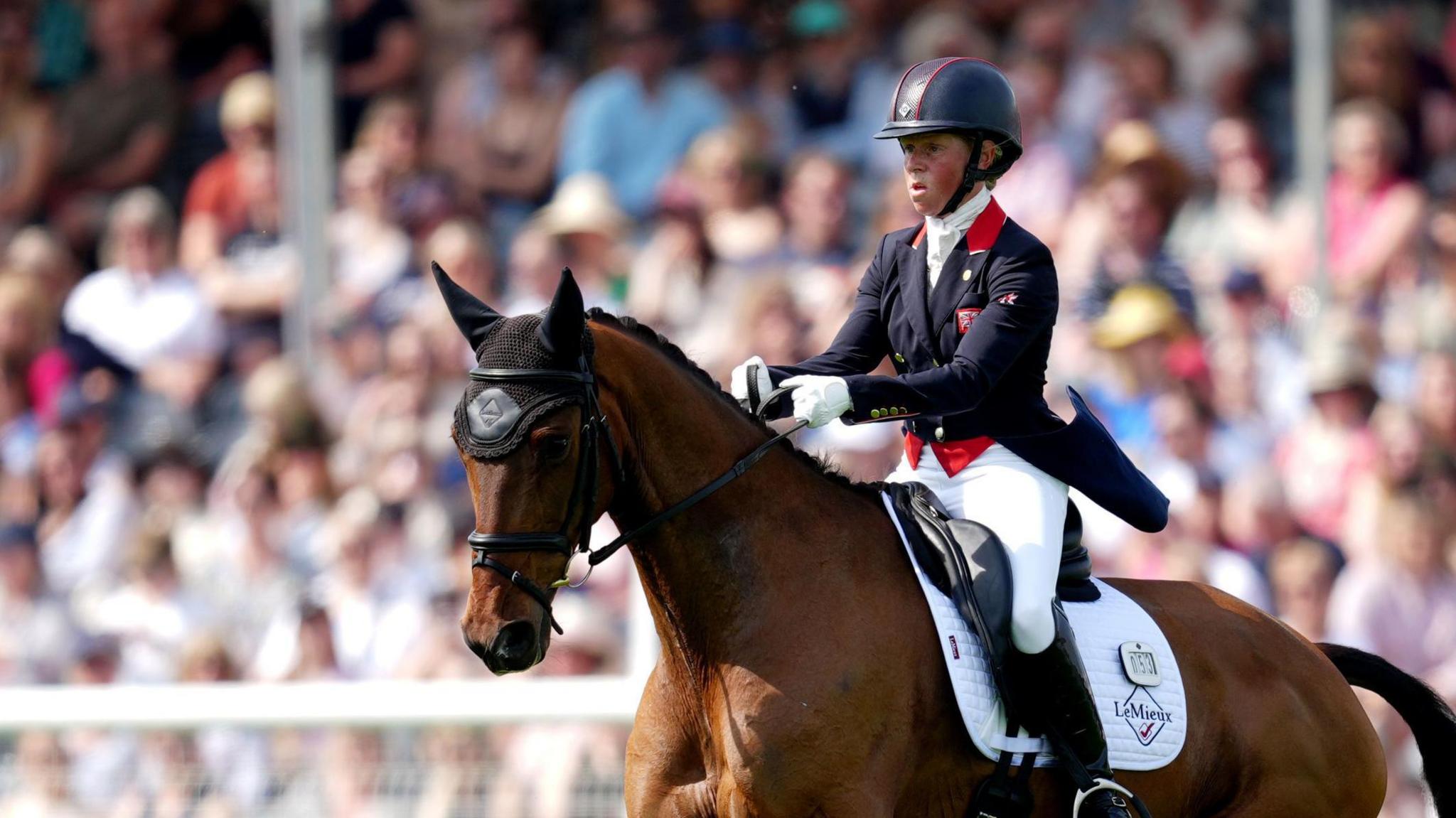 Ros Canter, dressed in blue and white equestrian clothes and a dark blue helmet, rides a brown horse in front of a crowd at the Badminton Horse Trials 2024.