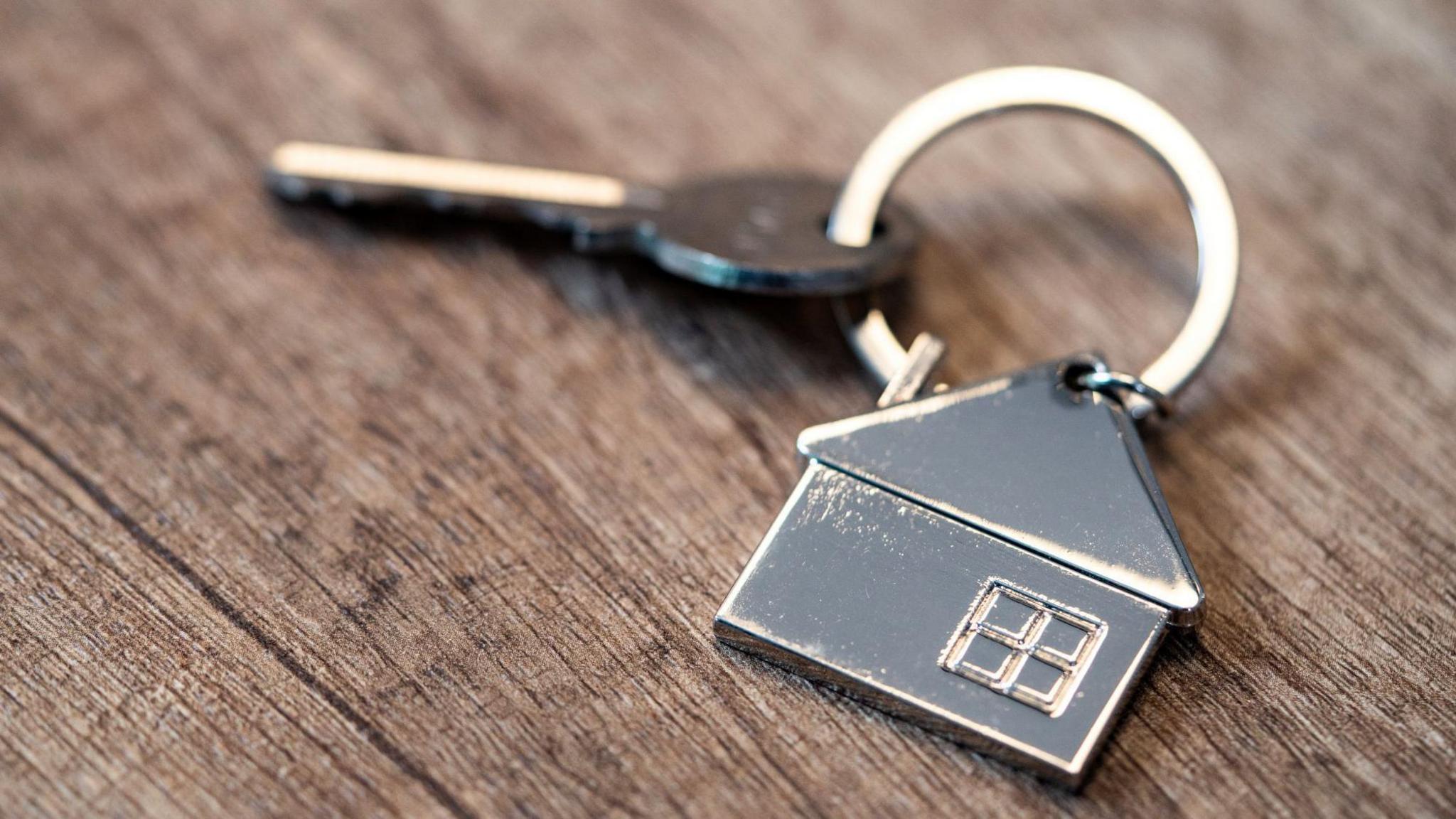 A silver-coloured house key attached to a house-shaped key ring. The items are laid on a wooden table top.