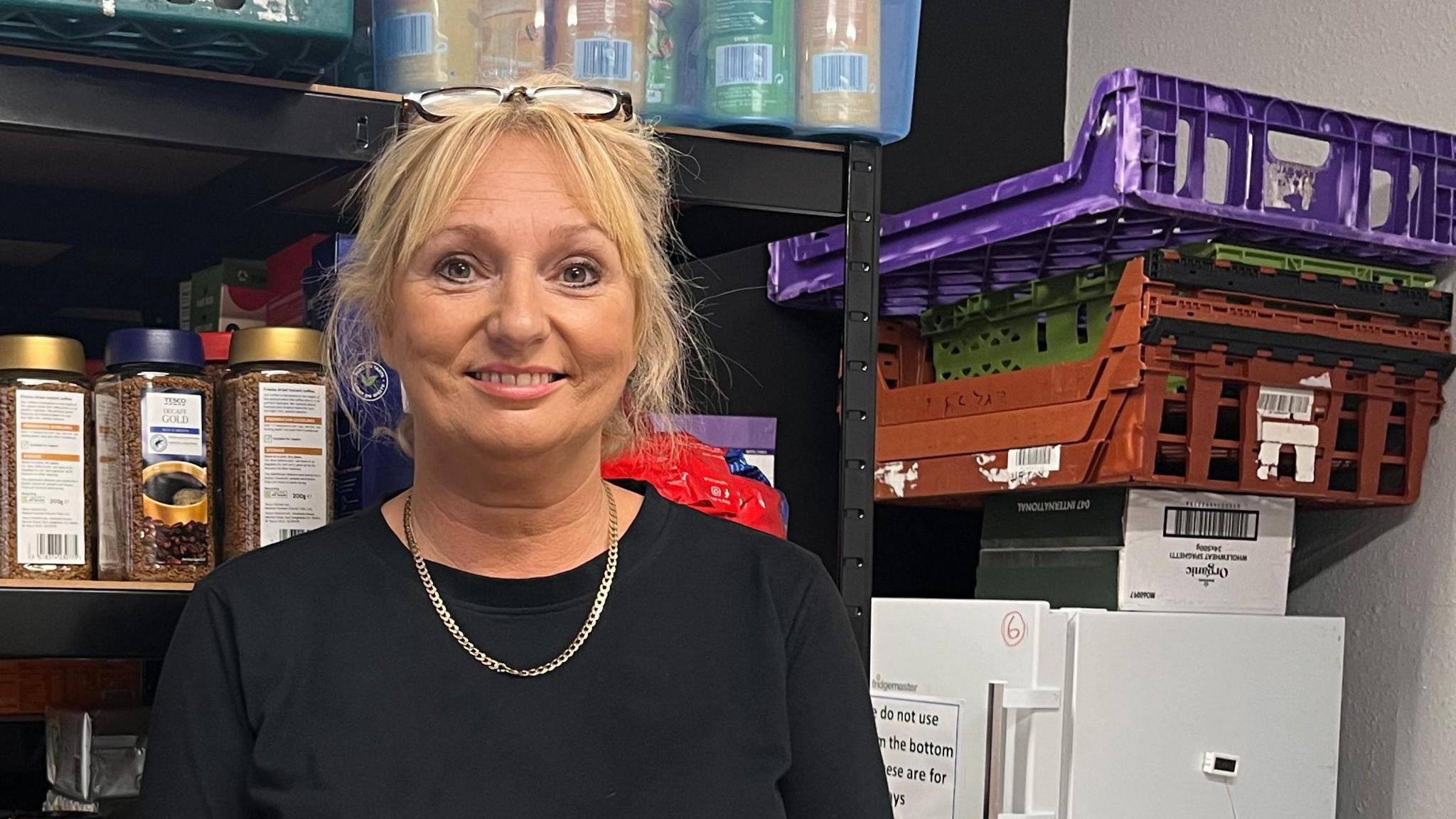 Jo Ramsay wears a black top, a gold necklace and glasses on her head. She has blond hair and is smiling at the camera. She is standing behind shelves of food and household products in a community centre food pantry. 