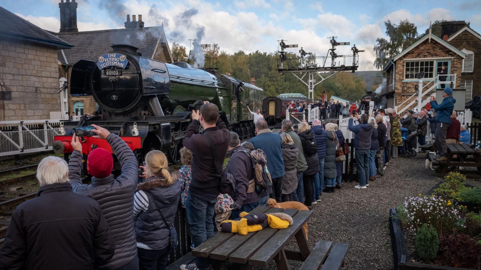 A crowd of people, including families and railway enthusiasts, gather on a station platform to admire and photograph the historic steam locomotive "The Flying Scotsman," numbered 60103. The engine emits a plume of steam as it prepares for departure. The station features traditional stone buildings, a wooden fence, and a signal gantry.