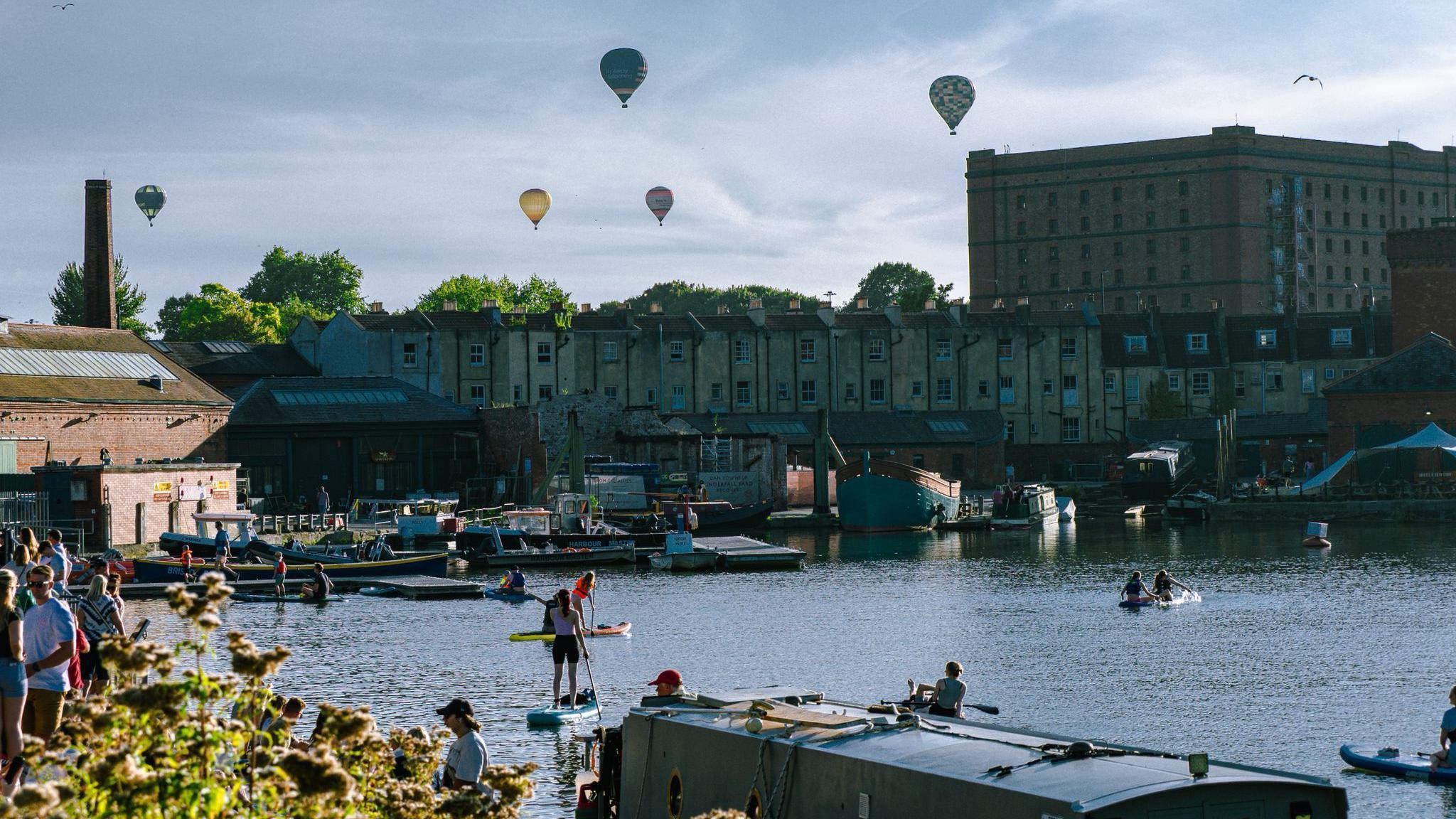 Hot air balloons are flying in the sky as people enjoy themselves in the summer sun on Bristol's harbour. Some are paddleboarding and kayaking while others stand alongside the water. The sky is blue and the sun is reflecting off the water.