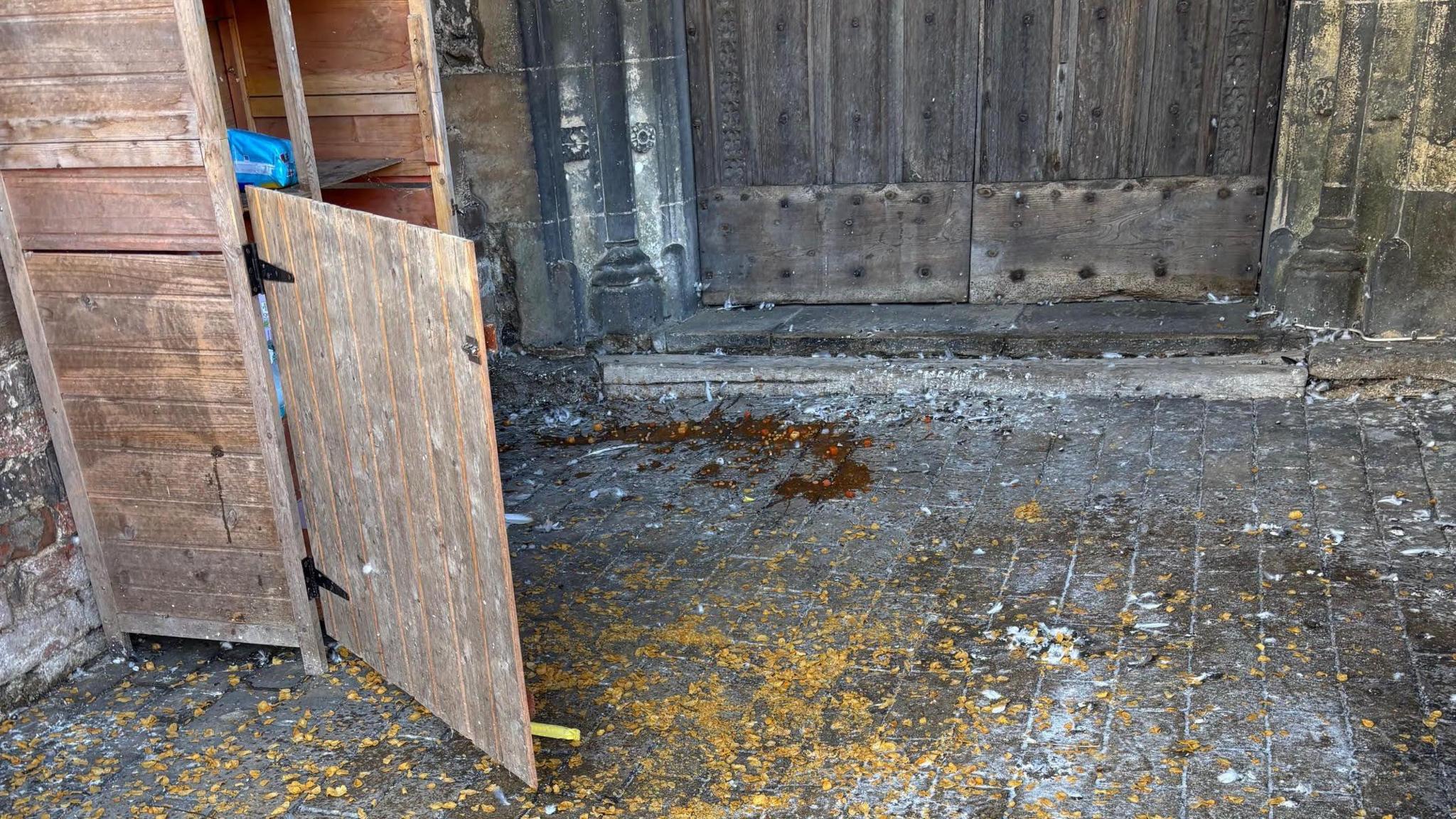 The contents of tinned food strewn across the floor in front of a church and a wooden outdoor cupboard.