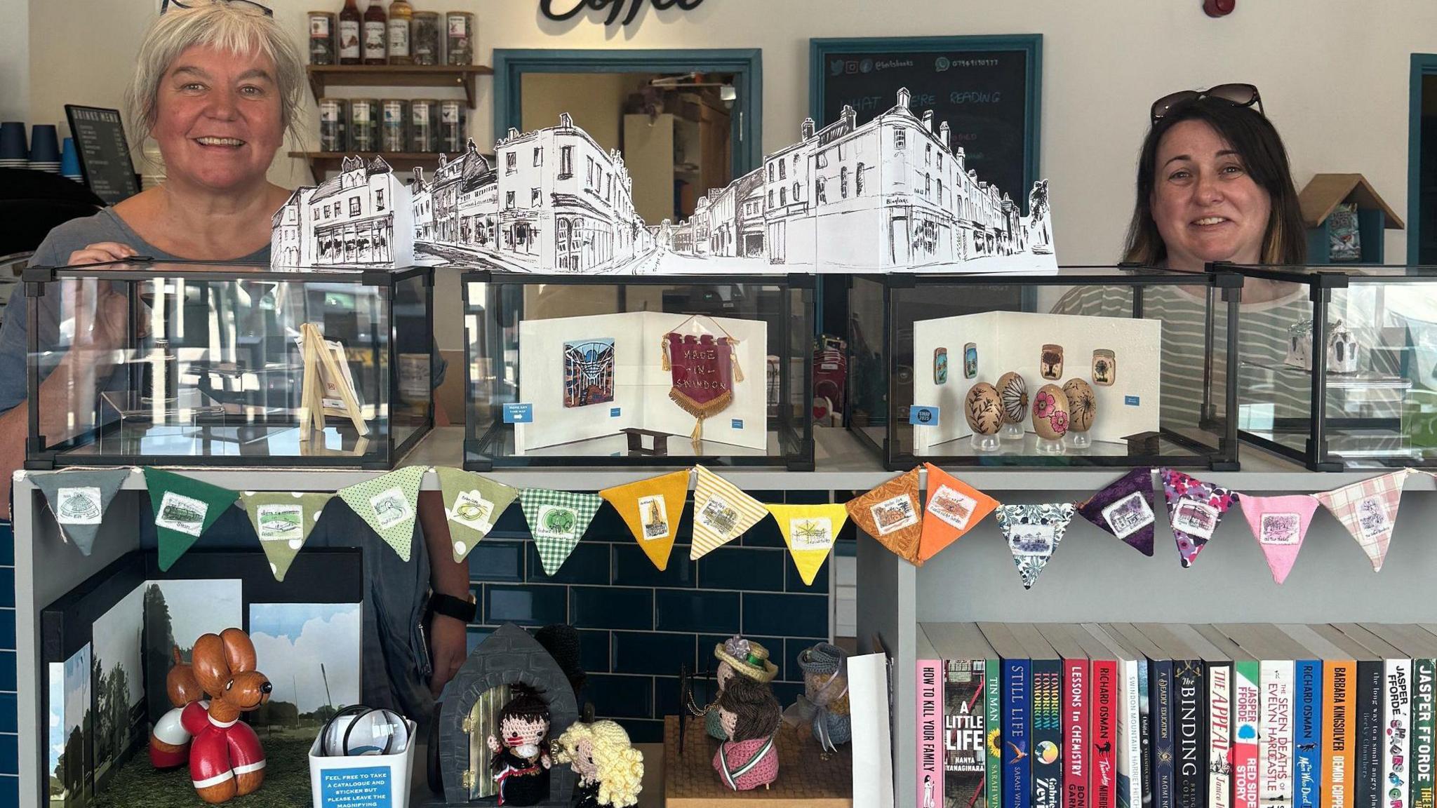 Two women stand behind shelves - on top is a row of clear shoeboxes with the displays in them. Bunting surrounds with a shelf underneath having knitted figures.