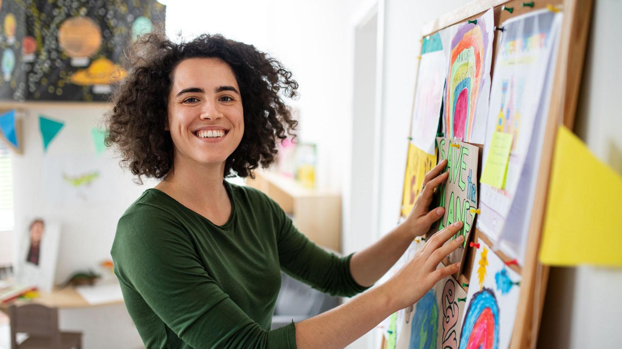 A female teacher smiling and putting things on a noticeboard.