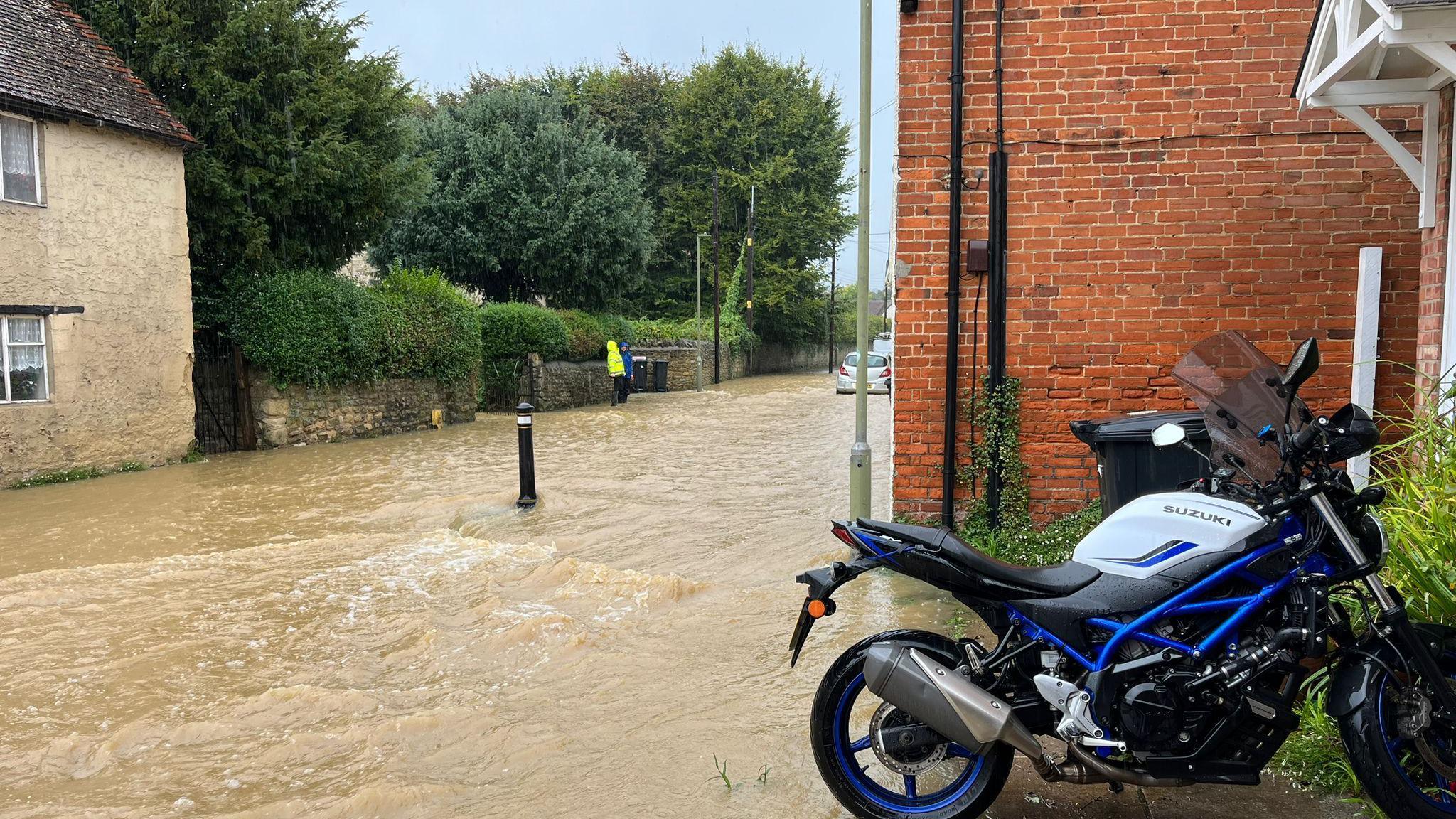 A motorbike in front of a house is next to a road that is flooded, with water clearly flowing down the street.