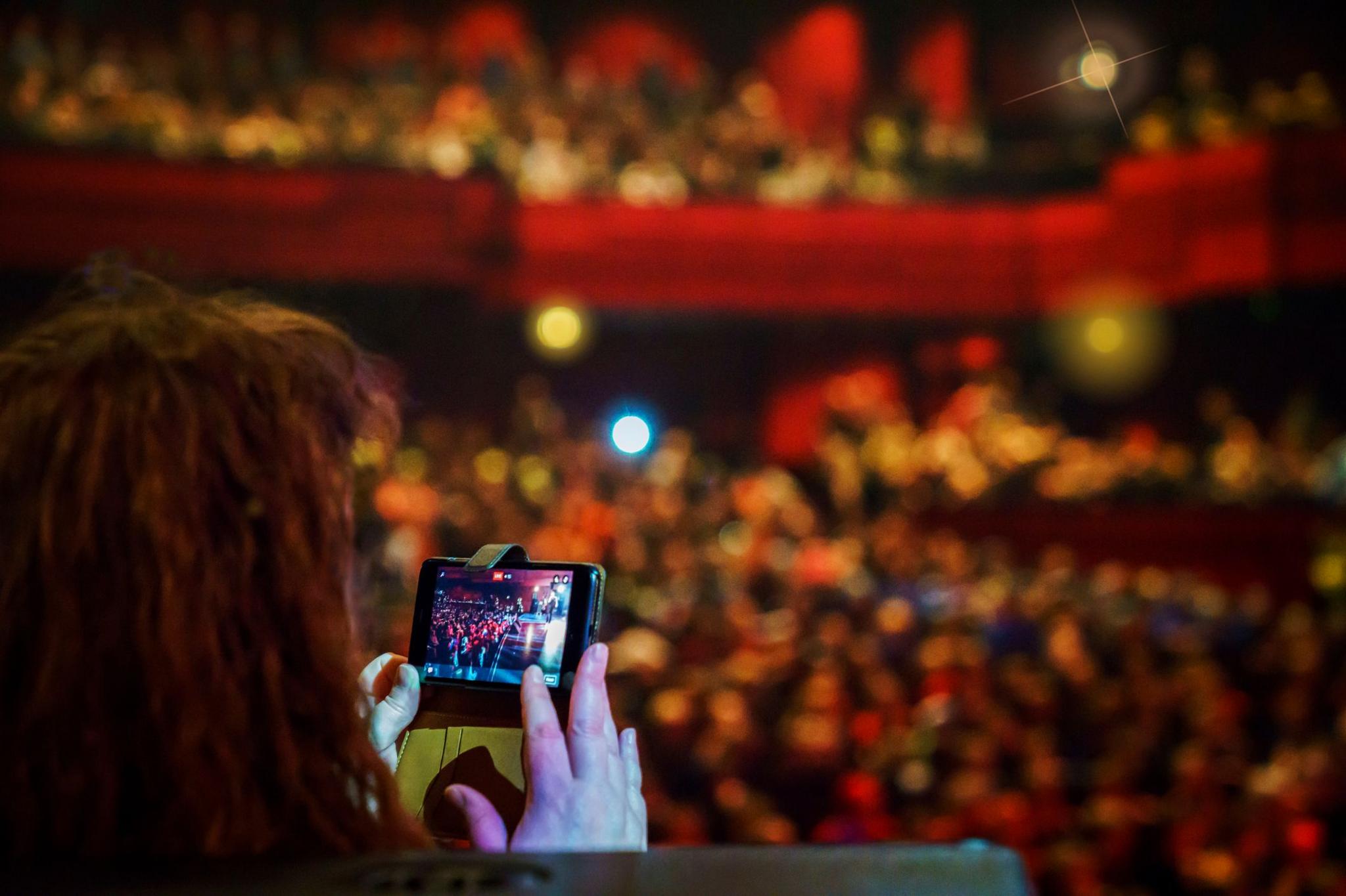 Back of a person's head can be seen as they hold up their phone to film the stage at the theatre 