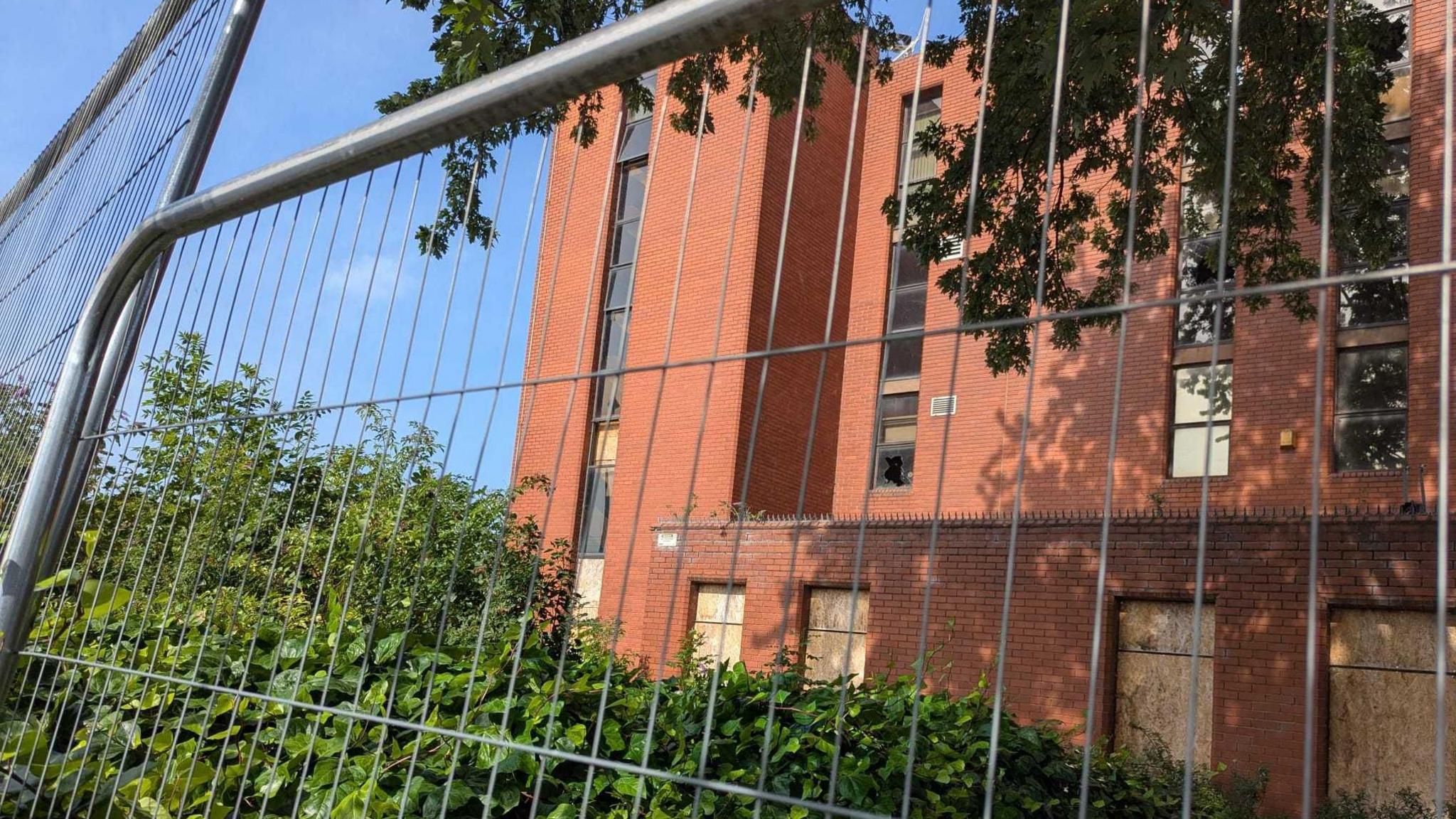 A red brick building behind fencing which has boarded up and smashed windows
