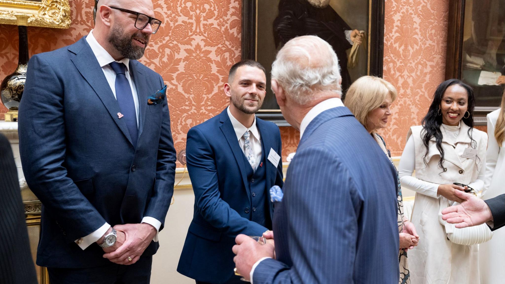 King Charles, in a blue pinstripe suit and his back to the camera, shakes hands with a man in a three-piece blue suit in a line-up at Buckingham Palace. The man is stood in front of pink wallpaper and wall paintings. Nex