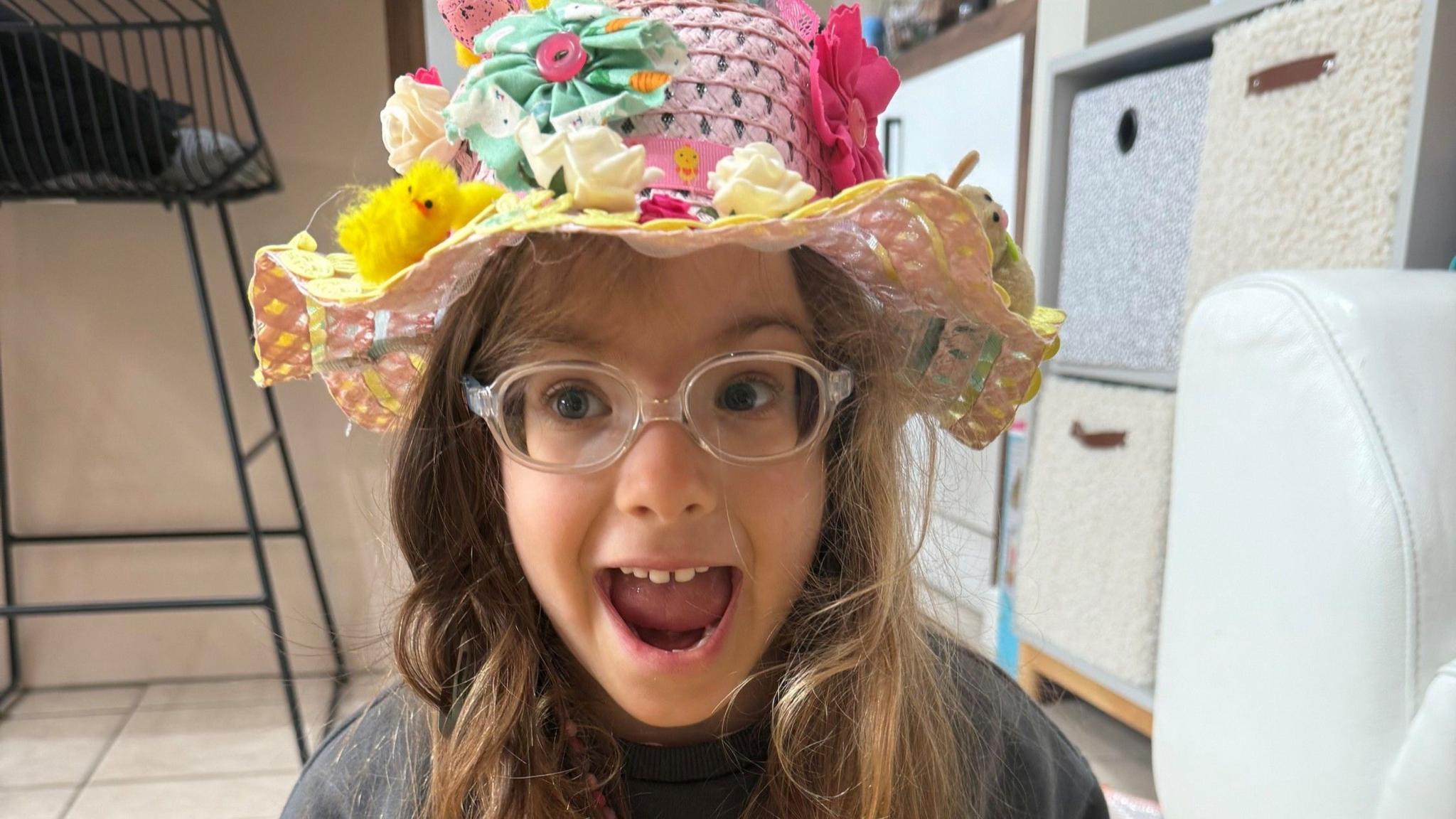 A young girl, with her mouth open, looking excited, wearing clear glasses, and an Easter hat with chicks, flowers and bright colours on her head. She is by boxes in a room with a chair to the left.
