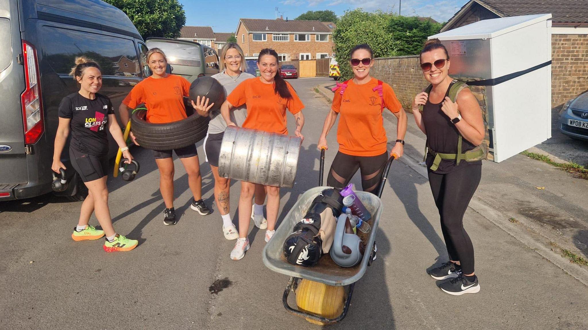 Commando Boot Camp members pictured in orange t-shirts, smiling for the camera carrying fridges, tyres and weights in their hands 