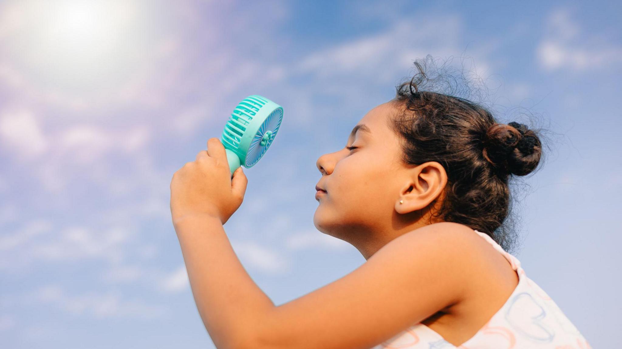 Close-up side profile of a young girl using a handheld portable fan outdoors under a bright blue sky during a hot summer day. 