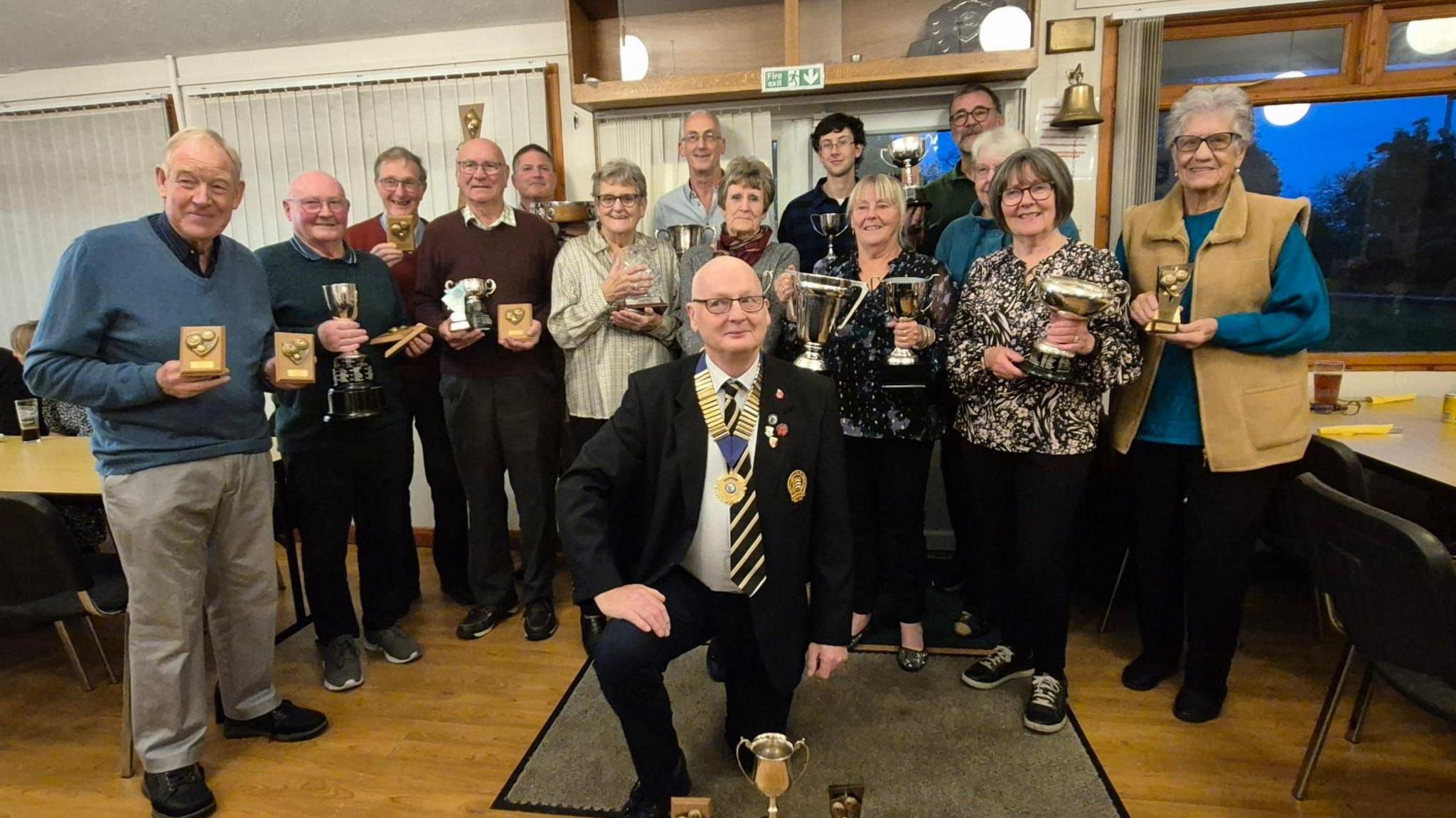 A group of people standing inside a room, side by side, holding trophies at a presentation ceremony. They are all smiling at the camera. One man is kneeling at the front of the picture. He is wearing a black suit and tie and a gold medal.