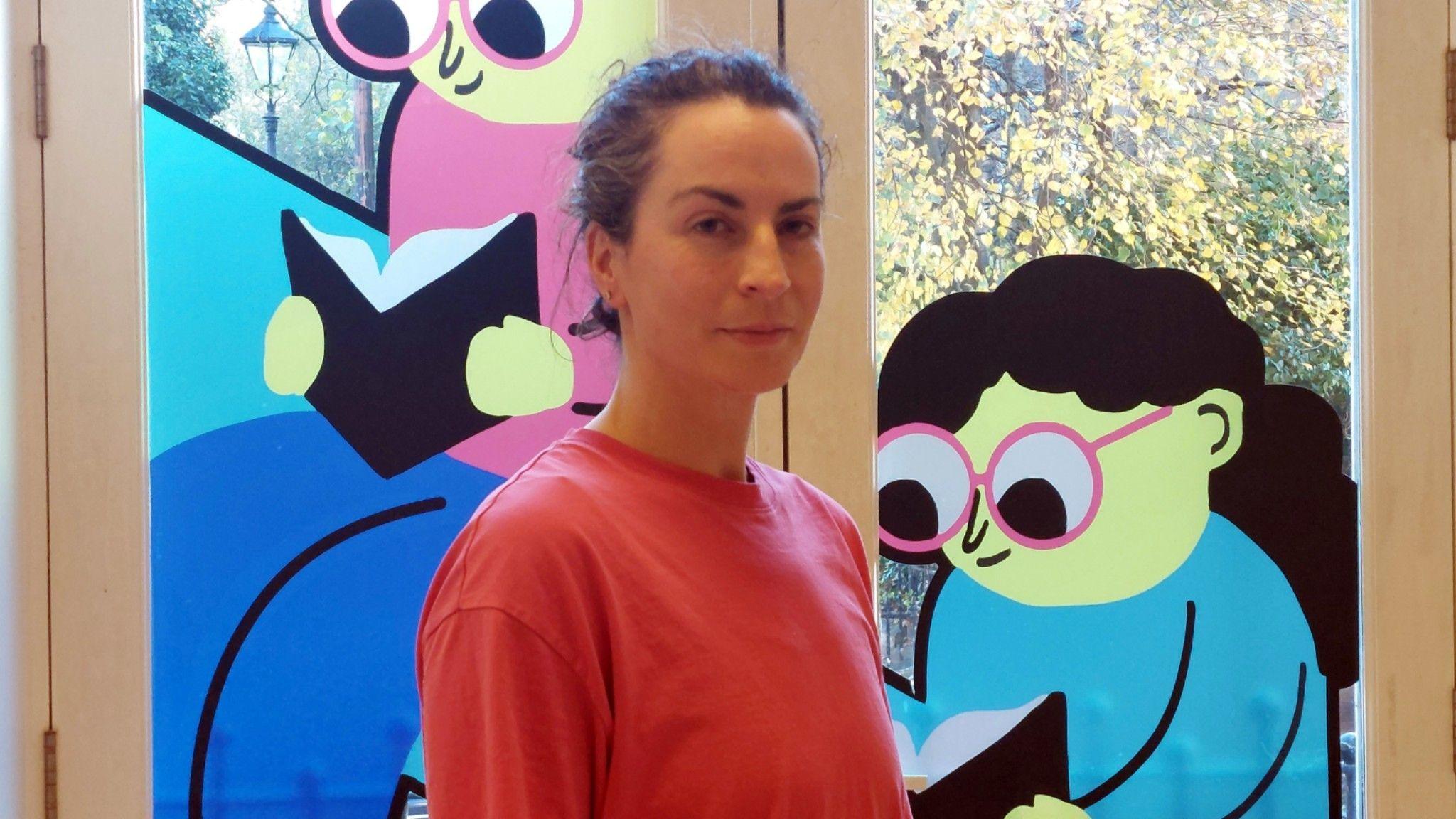 Helen Hall has brown hair and is wearing a pink top. She is standing indoors in front of large glass doors decorated with colorful illustrations of two cartoon figures reading books. Outside the glass, autumn trees with yellow leaves are visible.