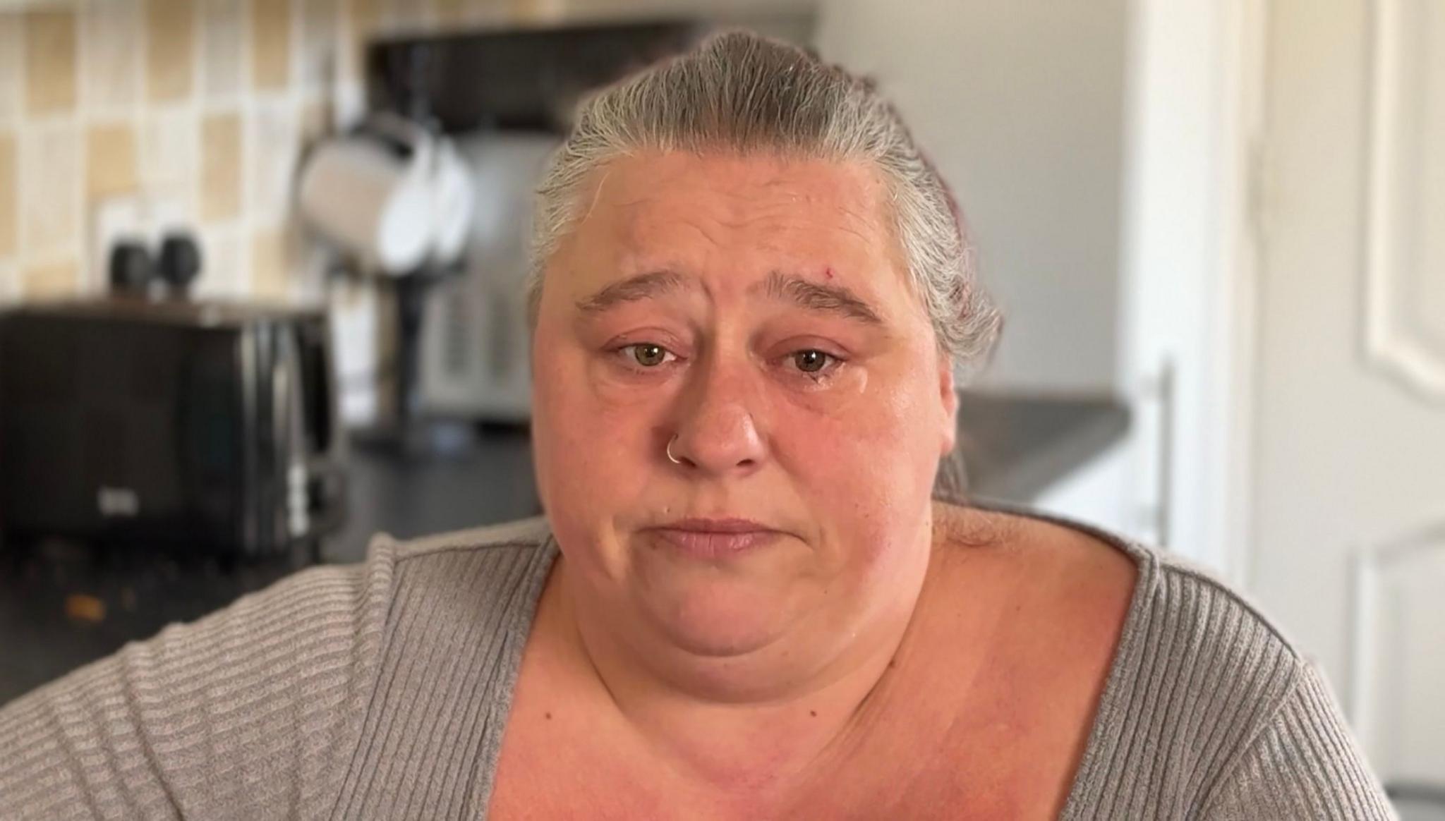 Headshot of a woman with tied-back, grey hair in a grey top looking into the camera with teary eyes.