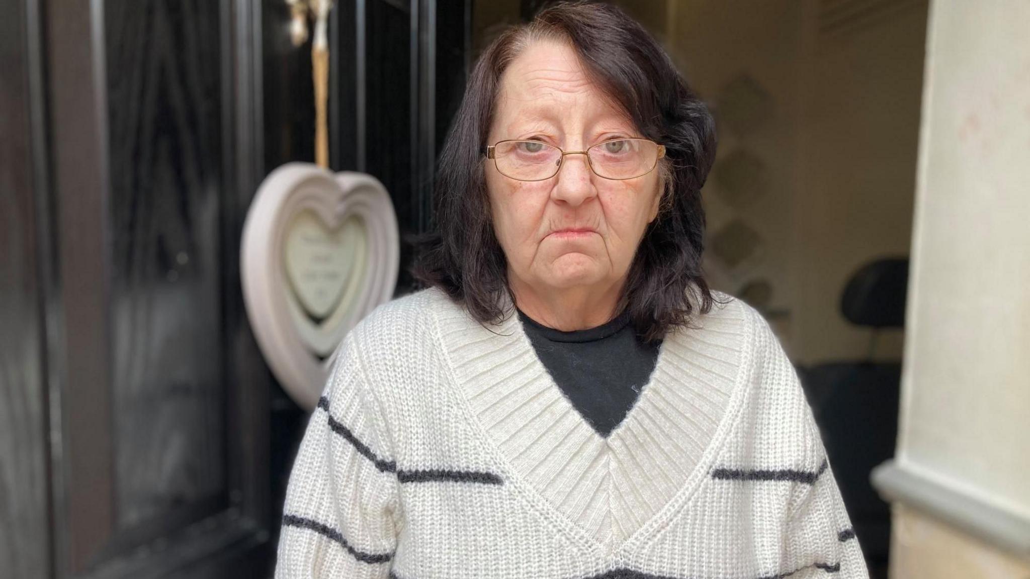 A woman with dark hair and a white jumper stands in the doorway of a property frowning at the camera