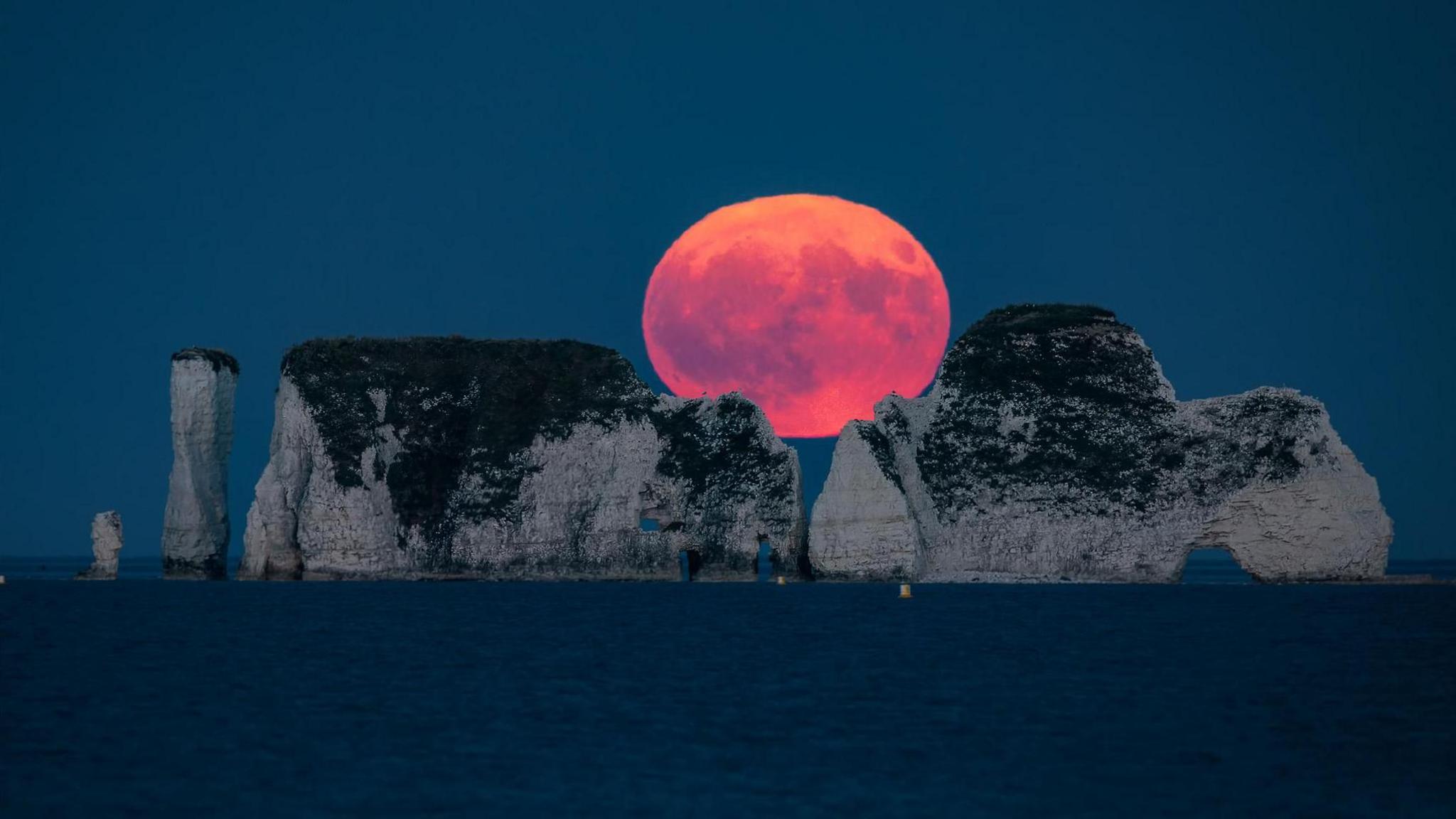 A huge-looking red/orange moon going down between two large cliff formations out to sea, with water in the foreground