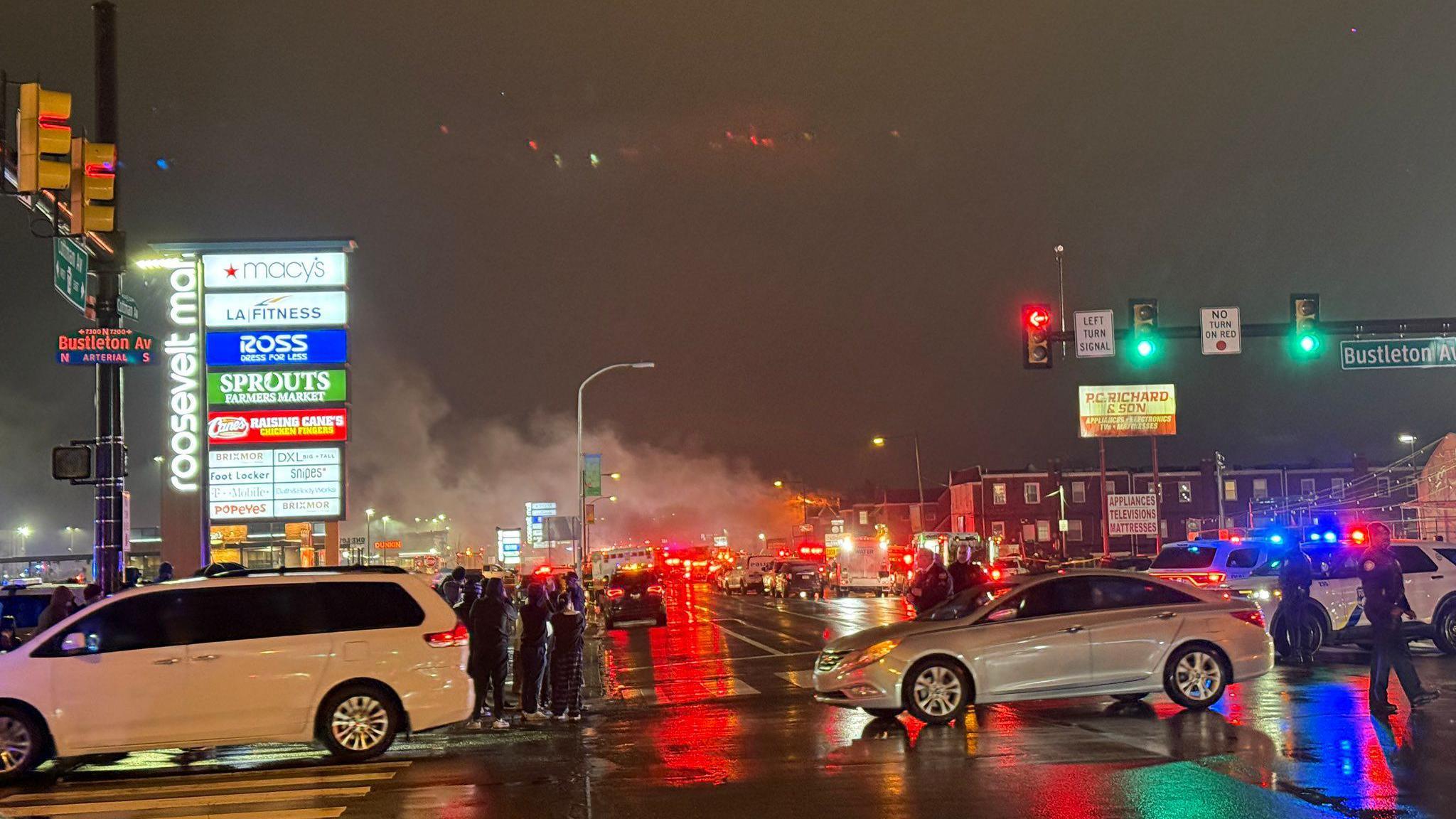 Smoke billows above a neighbourhood in Philadelphia. Cars are parked on the street and a crowd has gathered around an intersection. 