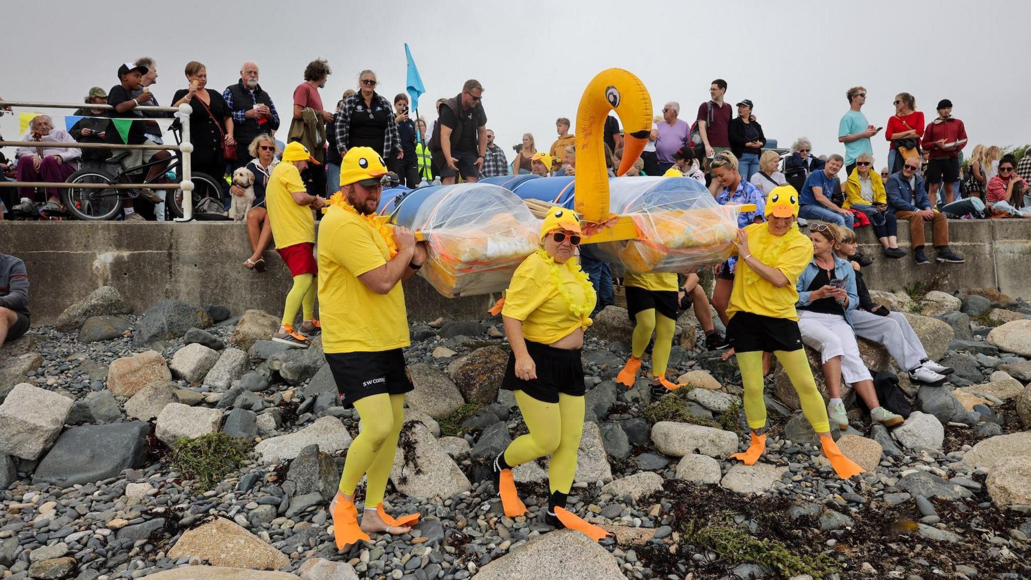 A team of six people dressed in yellow bird outfits brings a raft with blue barrels and a yellow bird's head down a rocky beach while spectators look on.