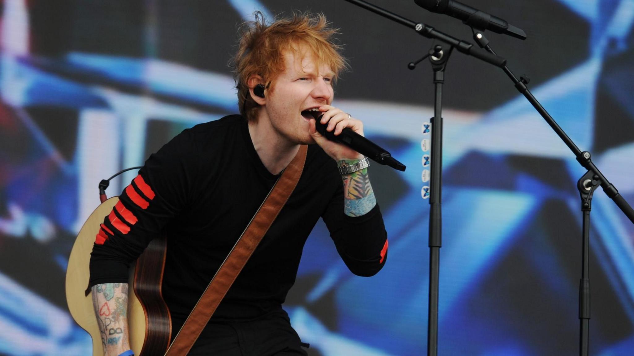Ed Sheeran on stage singing into a microphone and holding a brown semi-acoustic guitar.