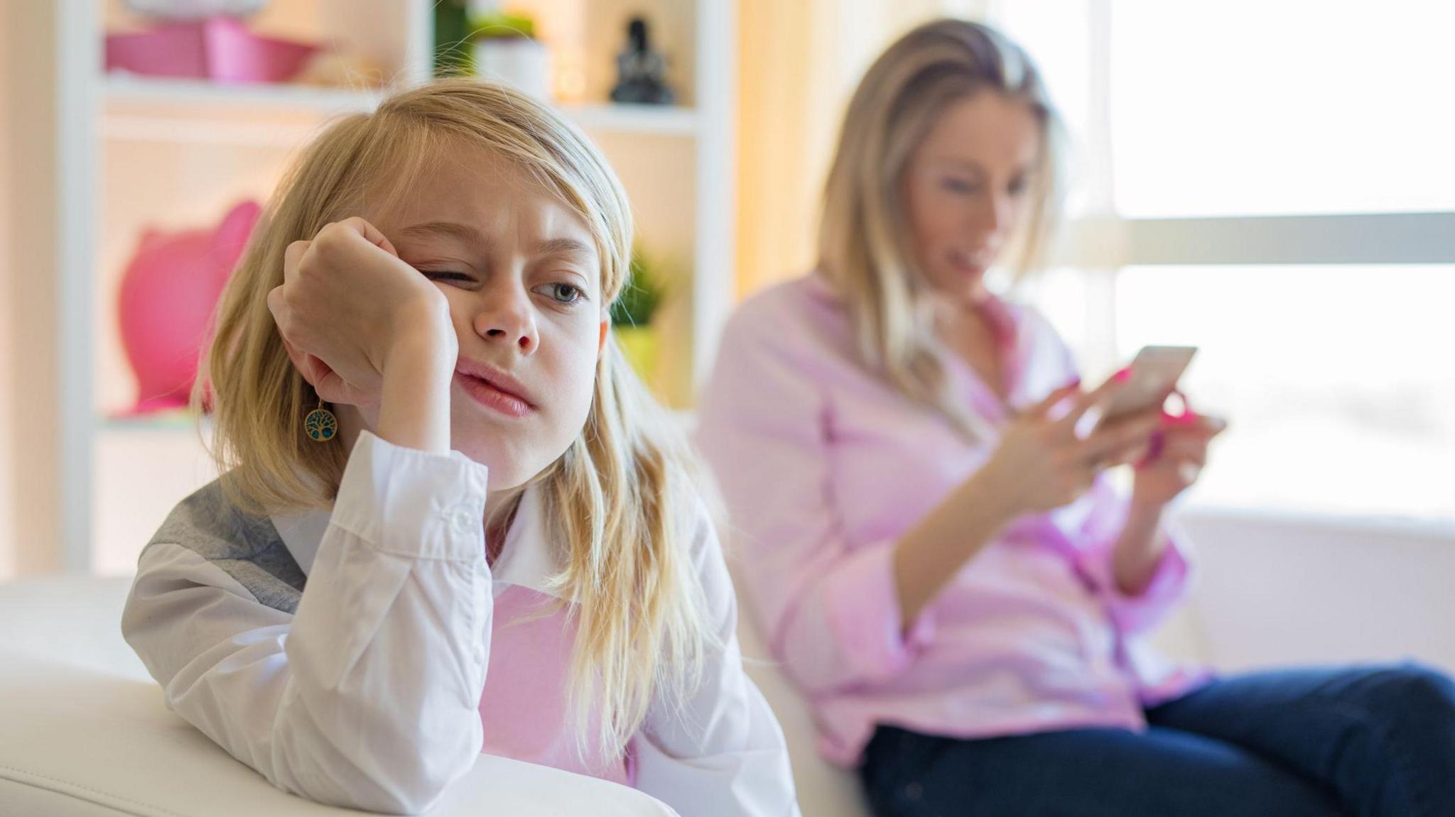 child looking bored while parent is on phone in the background.