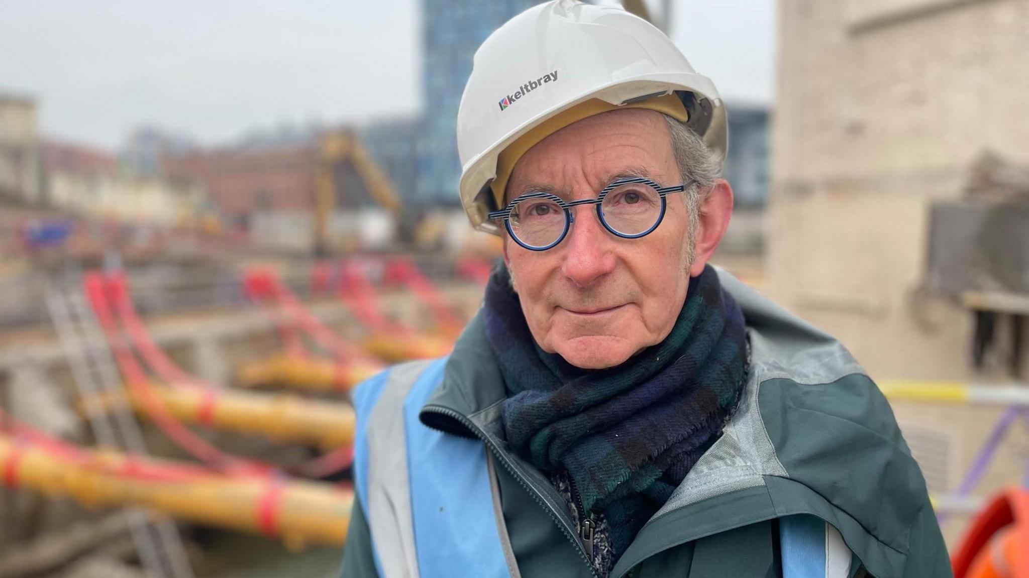 Simon Ogden, wearing a hard hat, scarf and winter coat, stands in front of a section of riverbank under construction