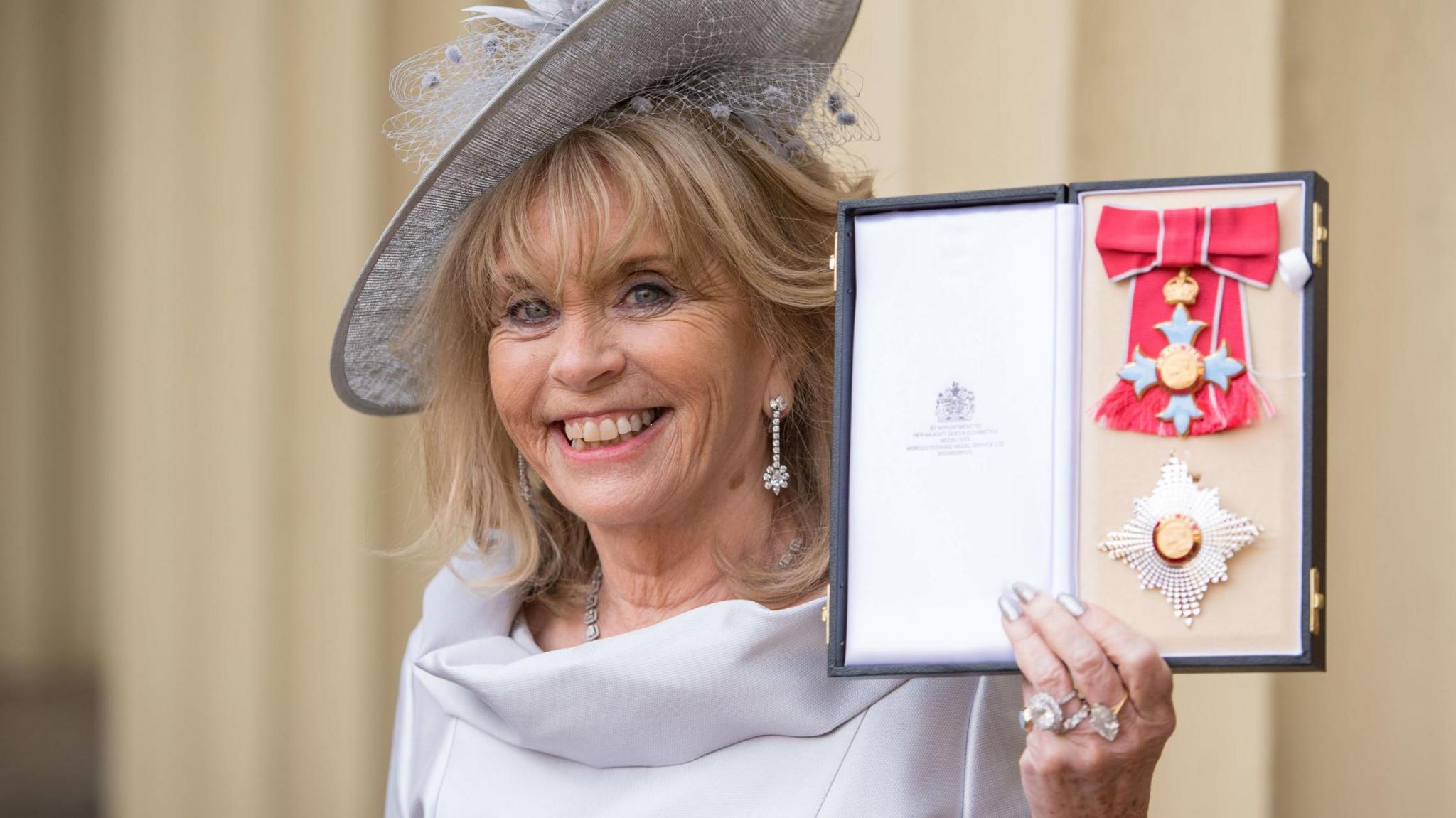 A smiling Dame Ann Gloag, wearing a white/grey hat, shows off her honours medal.