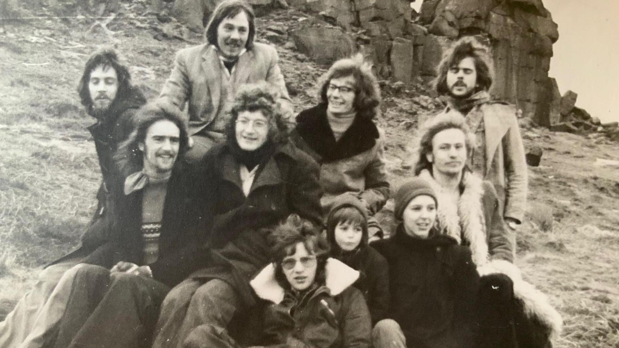 A black and white picture of a group pf people. They wear clothing which suggests the picture is from the 1970s. They are a mix of young men and women, with a couple of children also with them.