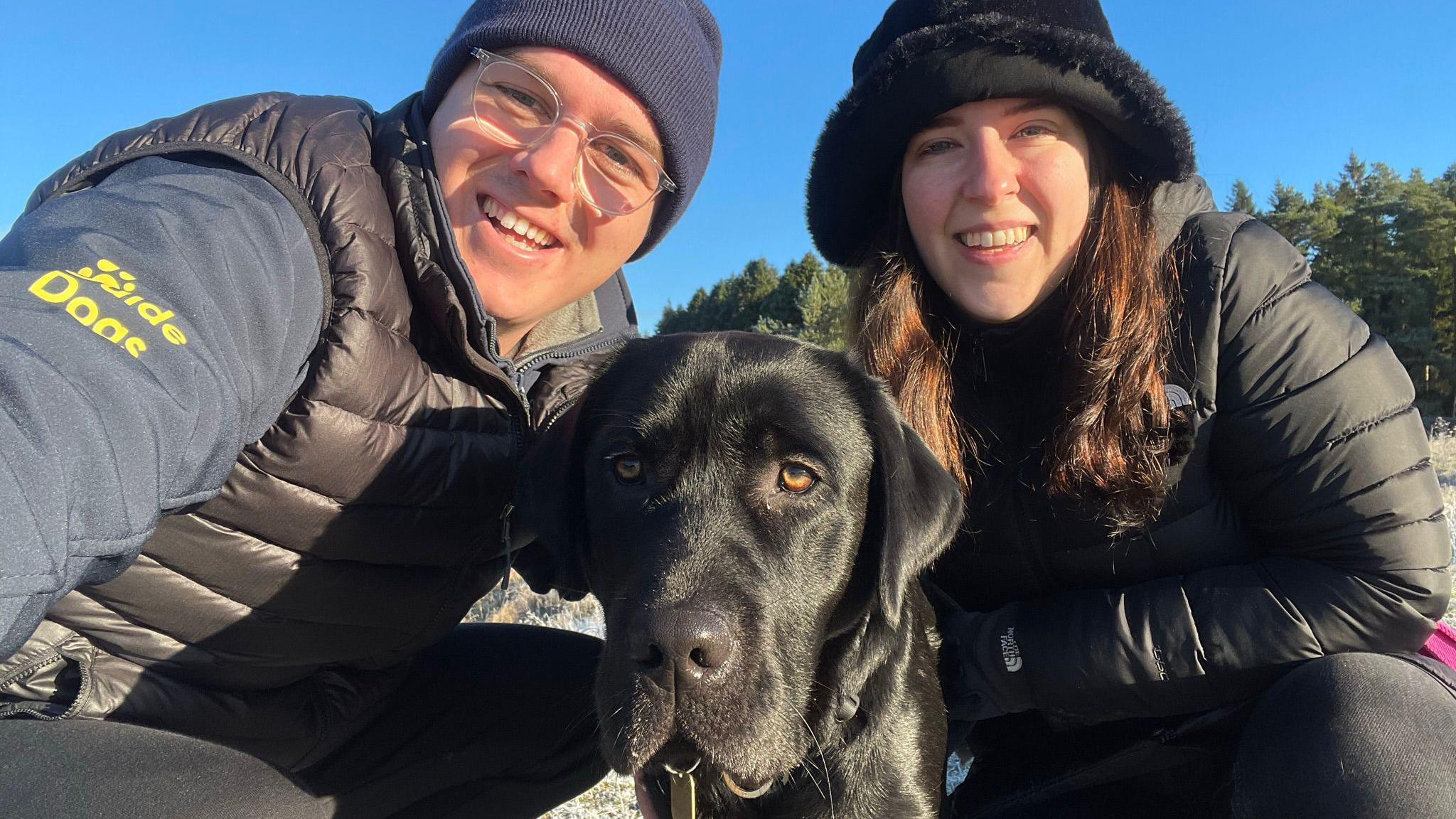 Adam Drysdale and his partner, Elle Manners. They are pictured outside on a dog walk. Adam is wearing glasses, a navy blue beanie and a navy blue jacket with the yellow guide dogs logo on the arm. Elle is wearing a black fluffy hat and a black coat. The couple are crouched down, smiling at the camera, with a black labrador guide dog with them.