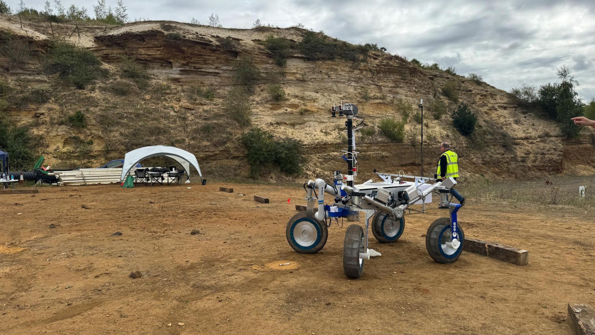 A four wheeled Mars rover is parked in a quarry with grey overcast skies overhead.