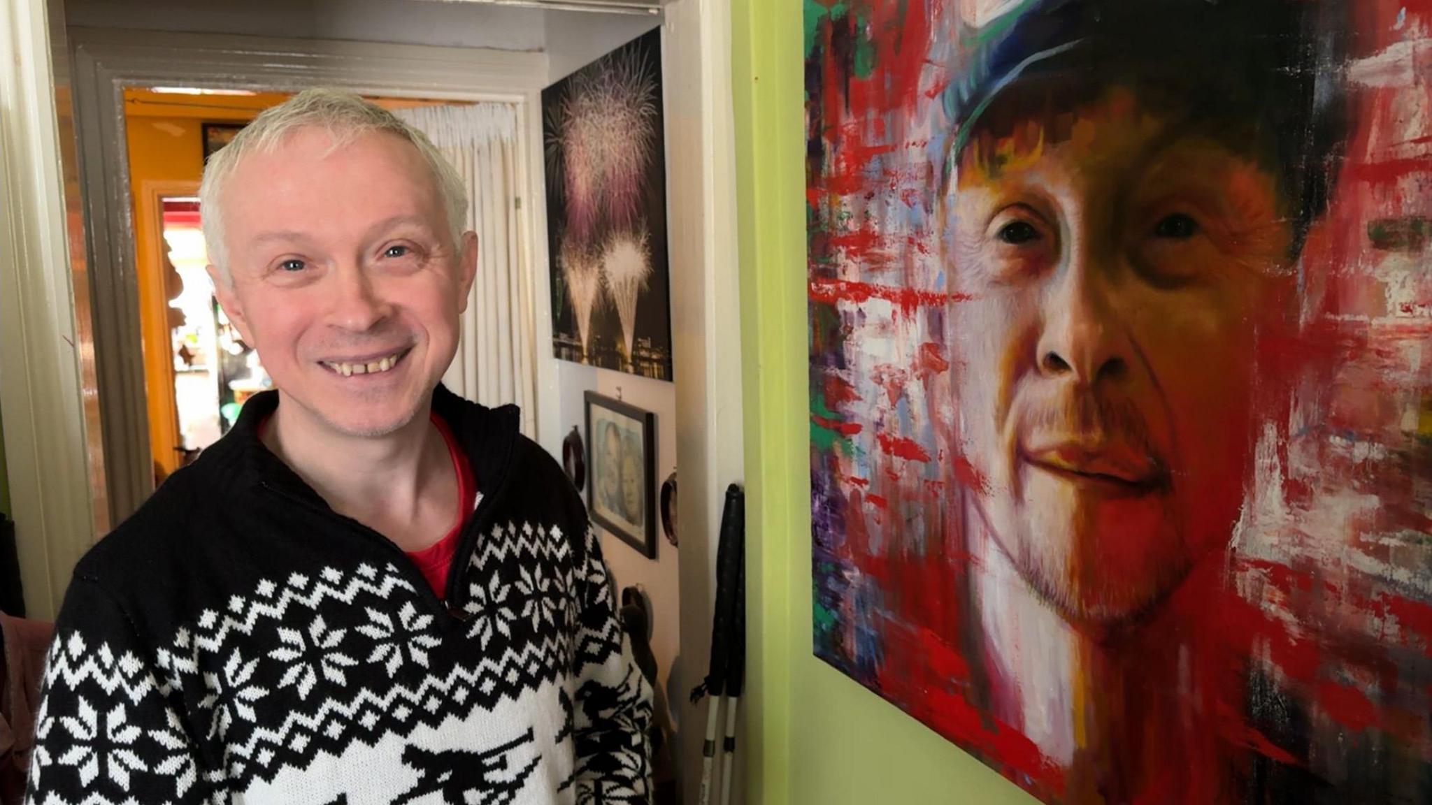 Clarke smiles into the camera and stands next to a portrait of him that is bordered by lashings of red paint. 