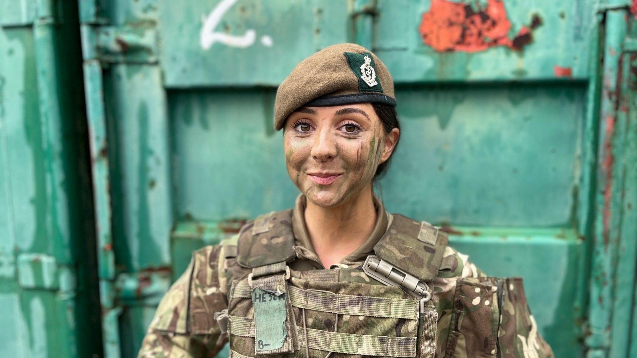 Rakela Kalemi wearing army fatigues, an Army beret and camouflage paint. She is standing in front of a copper green shipping container