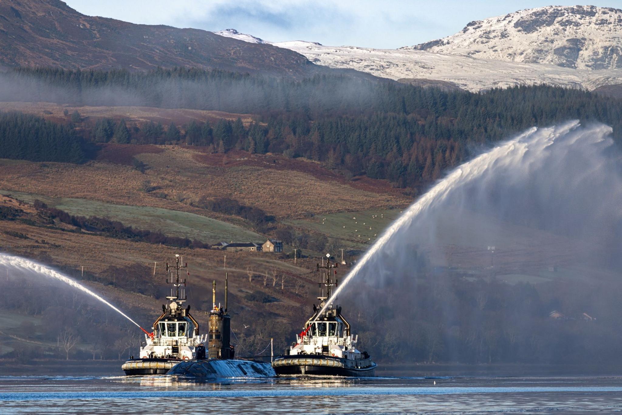 HMS Triumph submarine  at sea, with two tugboats sailing alongside it and firing their water cannons. Hills and trees are visible in the background. 