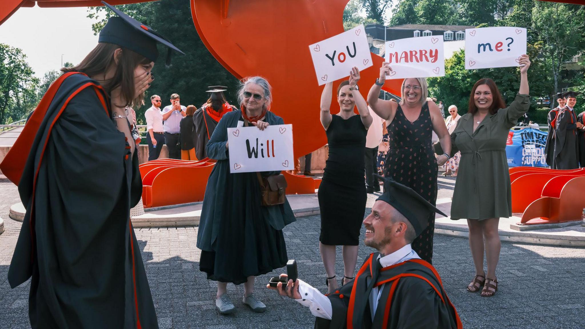 Kat standing with her hands over her mouth looking at Steve on one knee holding a ring. Four people stand behind holding signs saying 'Will you marry me?'